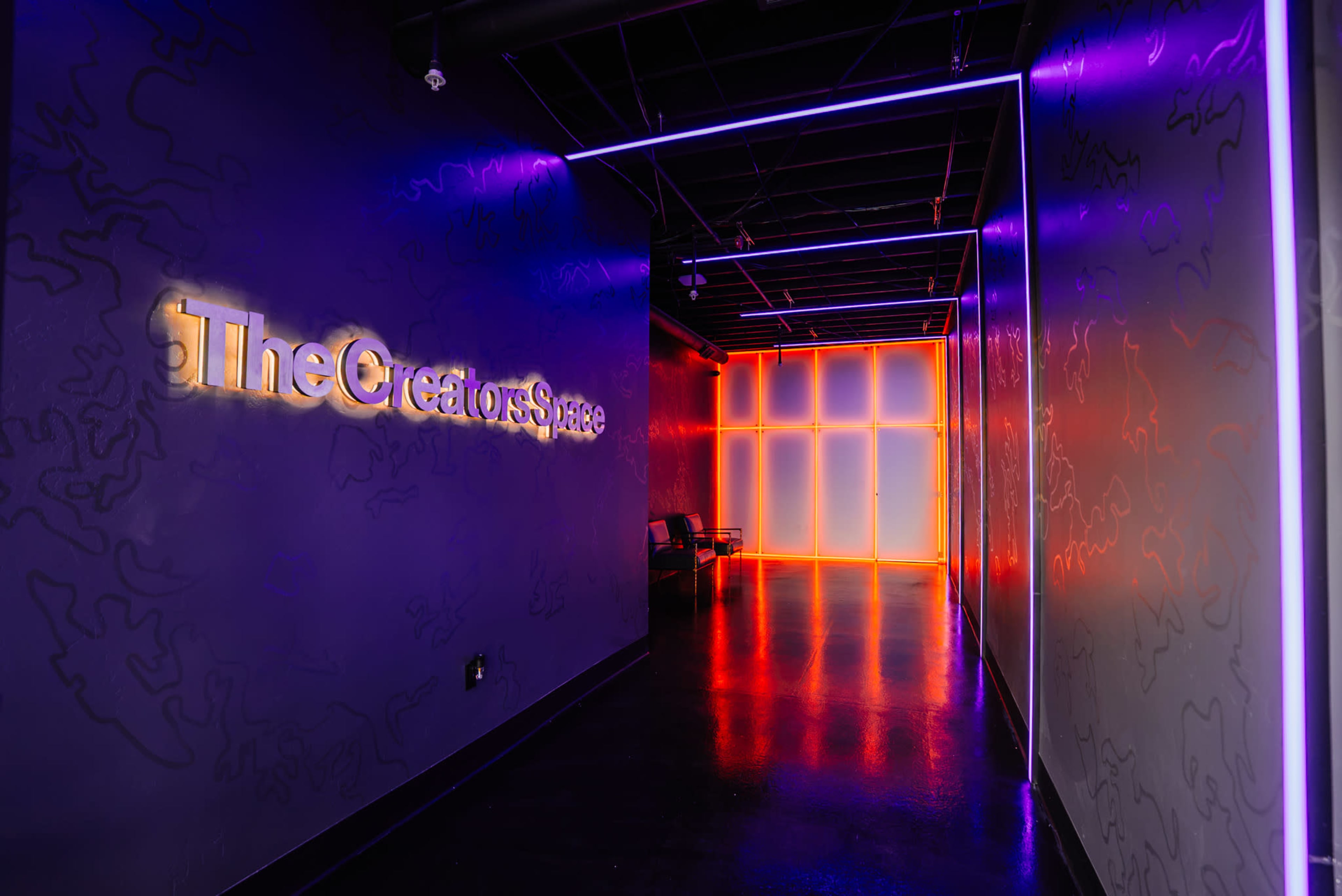 The image shows a modern hallway featuring illuminated walls with purple and orange neon lights, leading to a brightly lit area at the end, with the text "TheCreatorsSpace" displayed prominently on one wall.