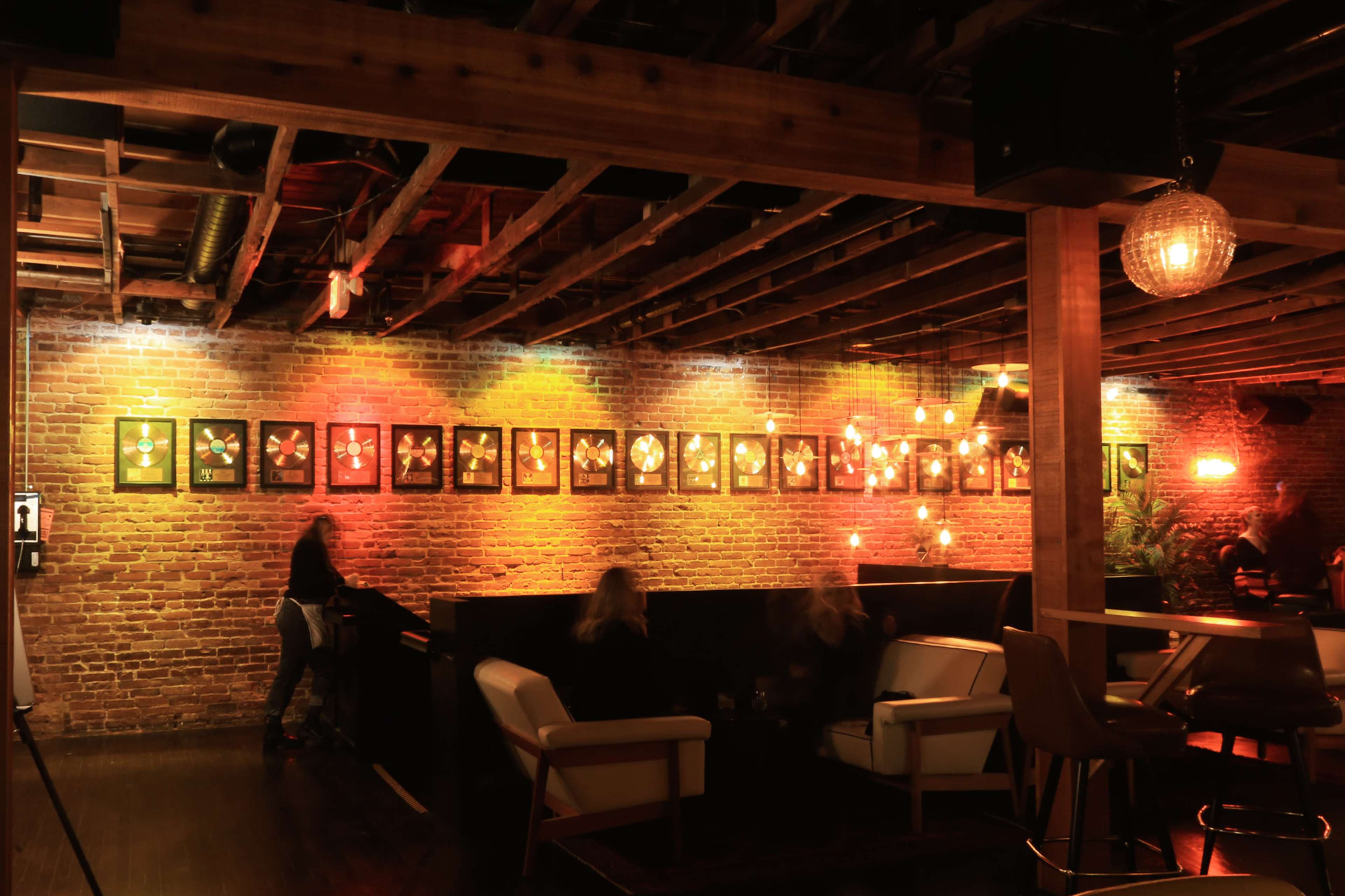 A dimly lit bar with a brick wall adorned with framed art and illuminated by pendant lights.