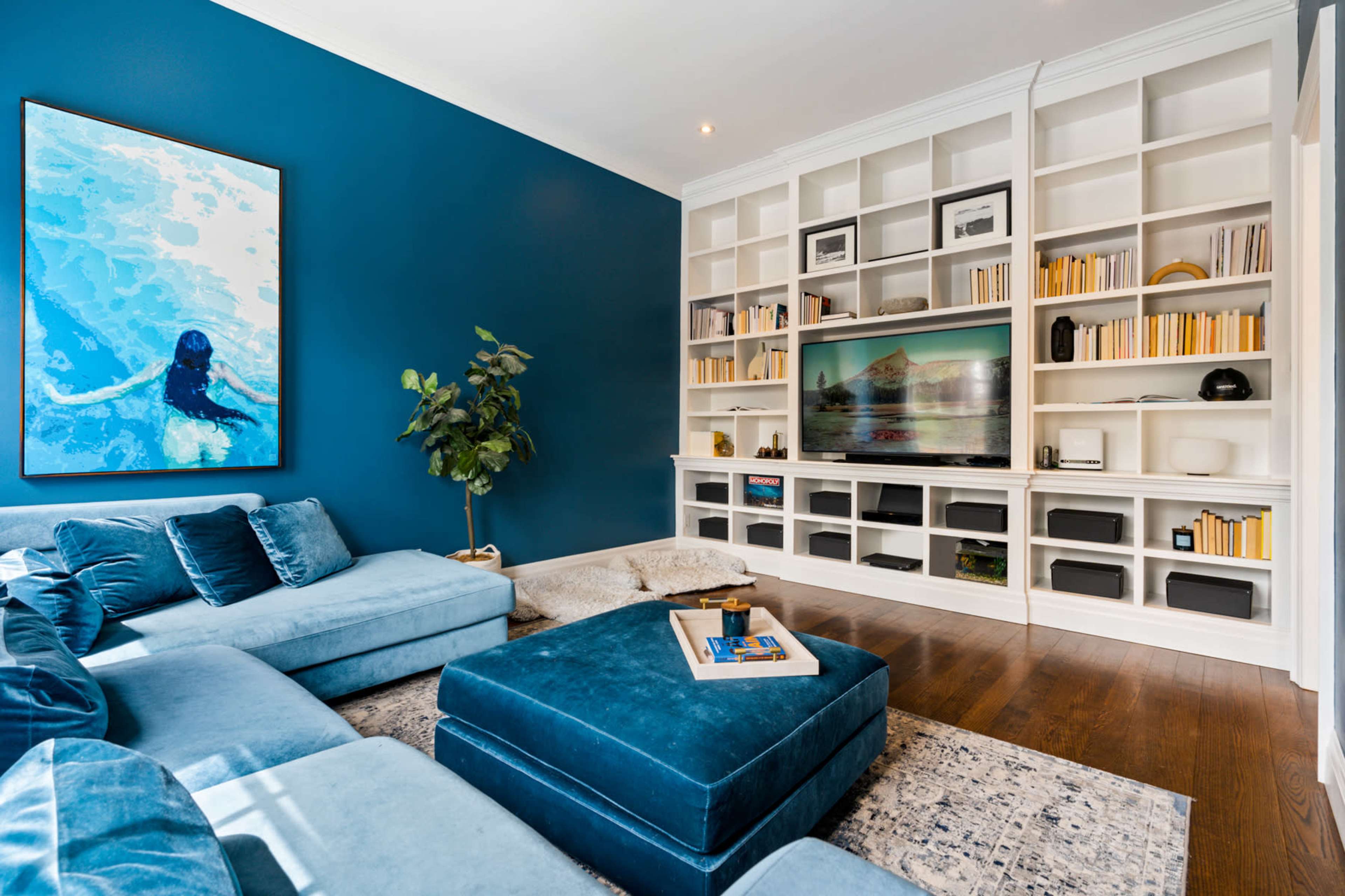 The living room features a blue accent wall, a plush sectional sofa, and built-in shelves displaying books and decorative items, with a large television in the center.