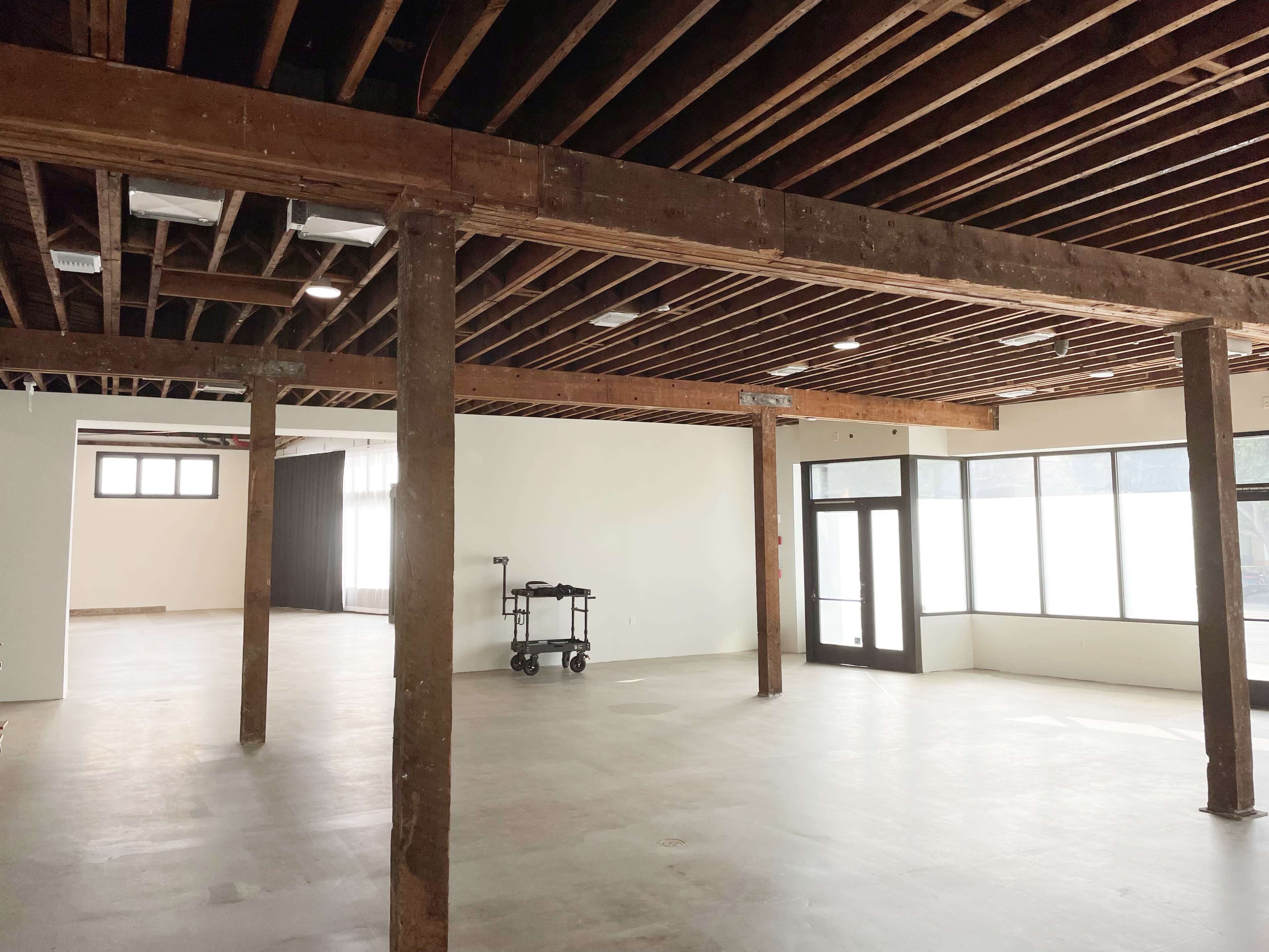An empty interior space with exposed wooden beams, concrete floors, and large windows along one wall.