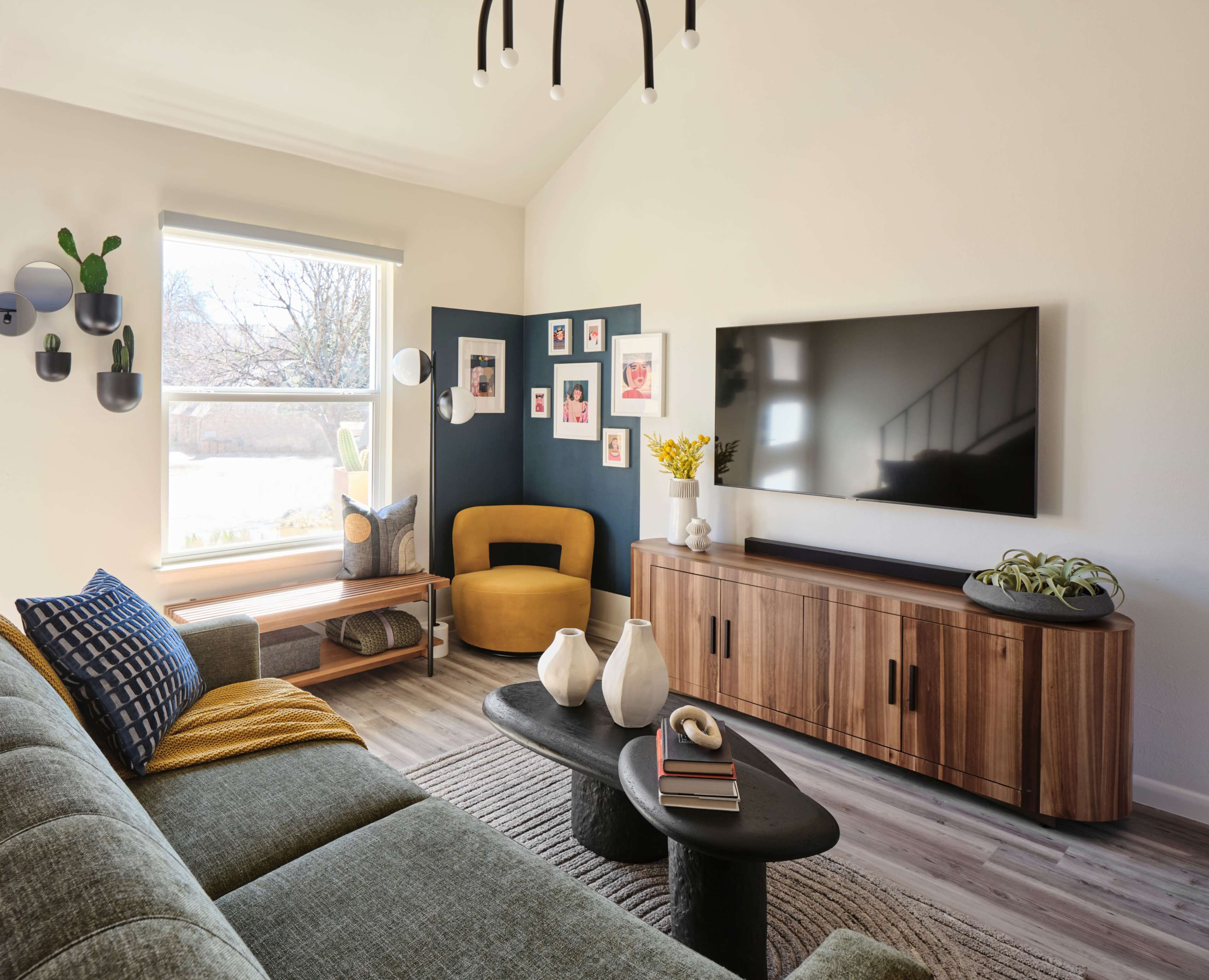 The image shows a modern living room featuring a sectional sofa, a round coffee table, a large flat-screen TV mounted on a wooden cabinet, and various decorative elements including plants and framed photos.
