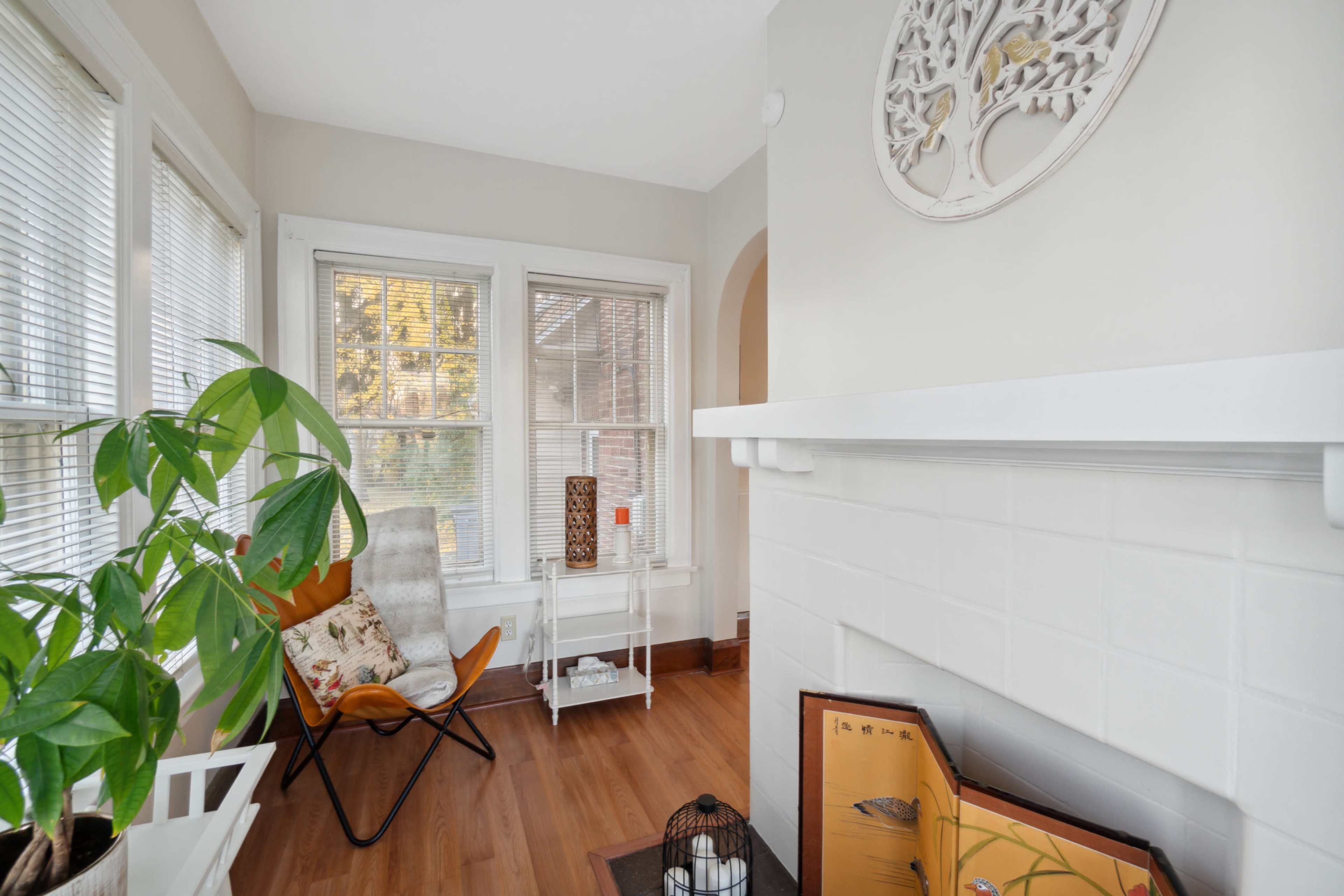 A cozy corner of a room with a modern chair, a potted plant, and a decorative wall piece, featuring a fireplace and natural light coming through the windows.