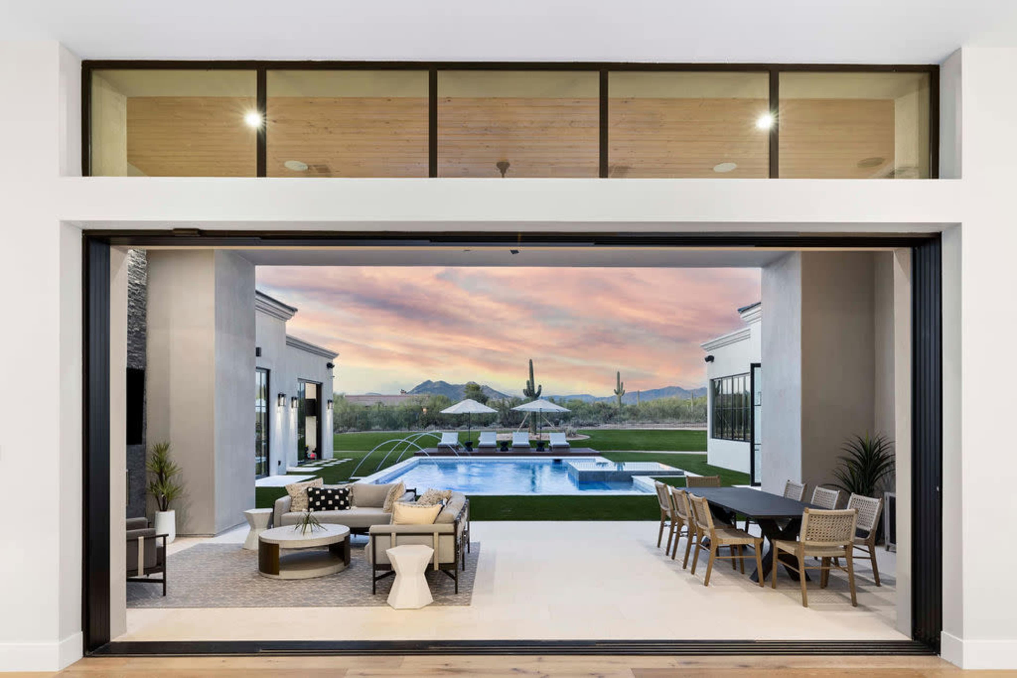 A luxurious outdoor area with a swimming pool, surrounded by lounge furniture and dining tables, set against a backdrop of mountains and a colorful sunset.