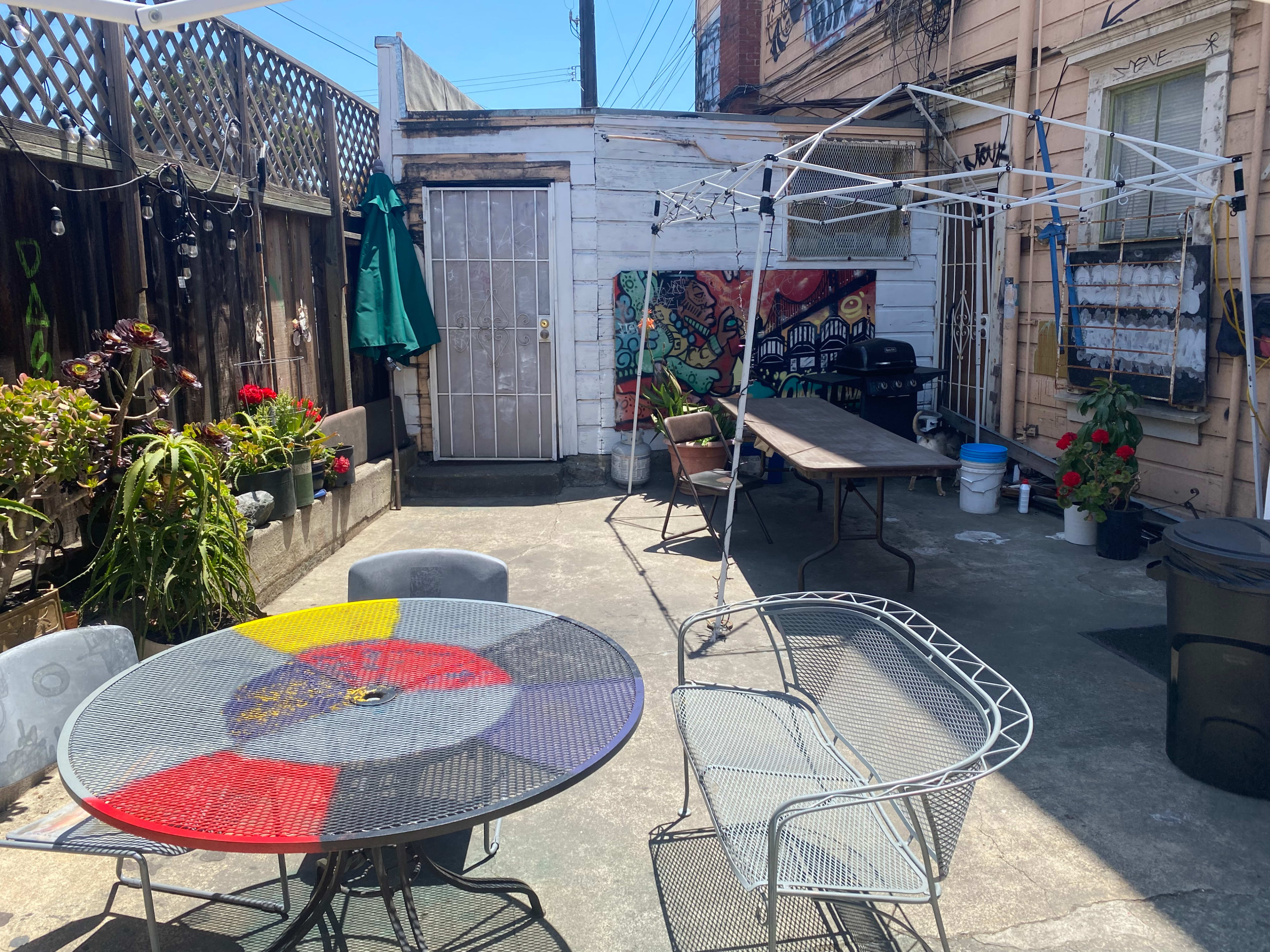 The image depicts a small outdoor patio area with a round table, several chairs, a weathered shed, and a canopy frame set up beside colorful murals and potted plants.