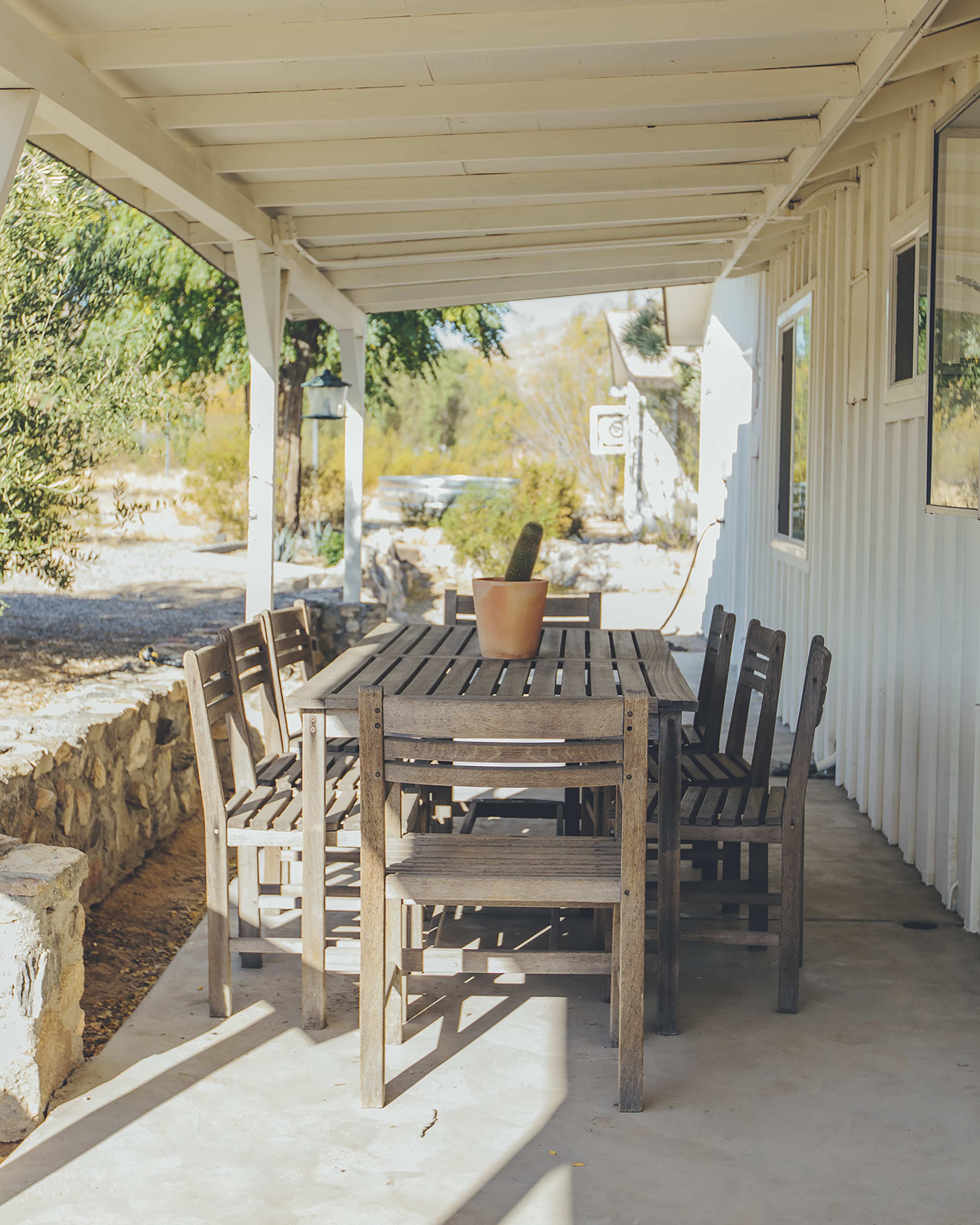 A wooden dining table with matching chairs sits under a covered porch, accompanied by a potted cactus.