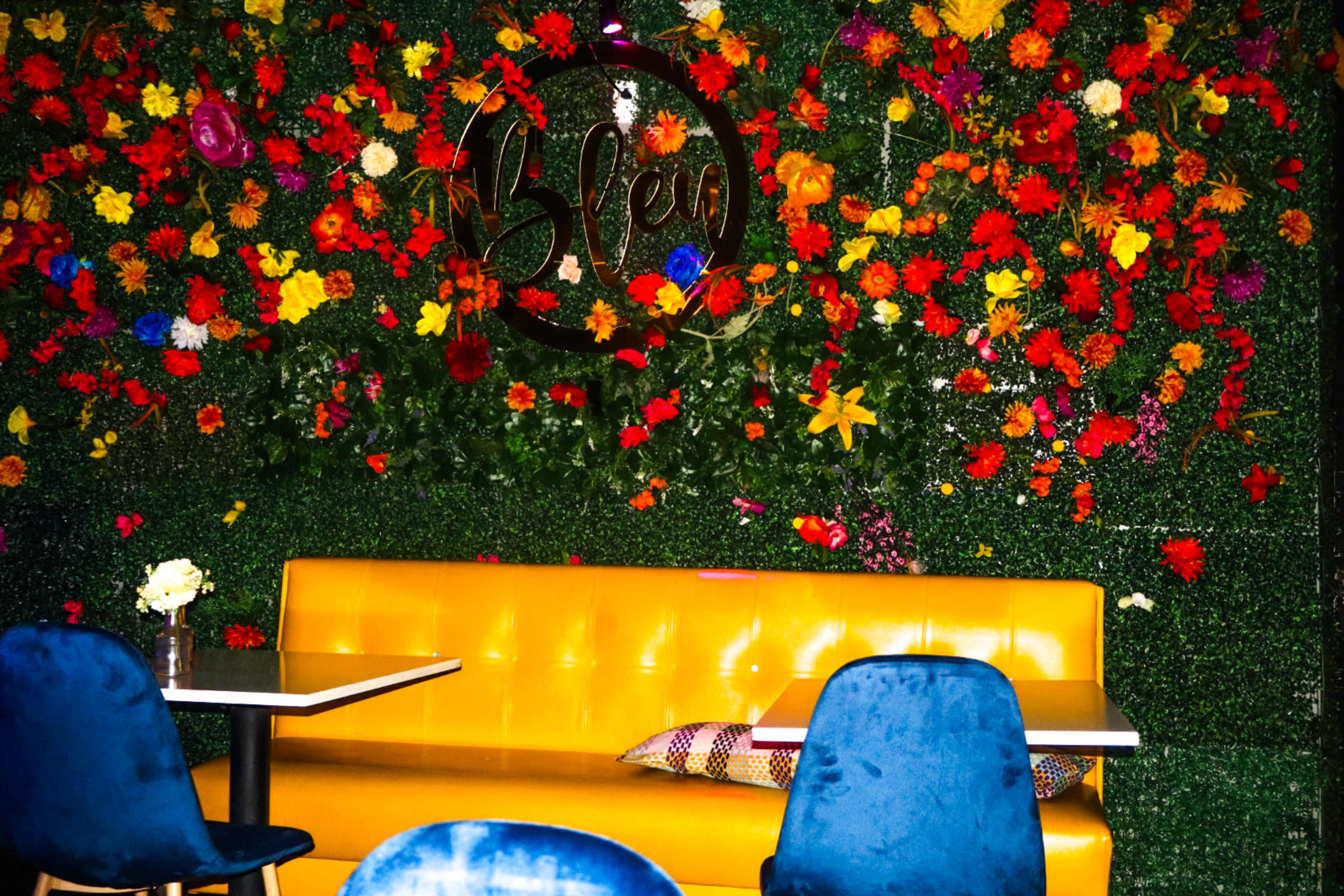 A vibrant wall of artificial flowers surrounds a yellow booth and tables in a modern café setting.