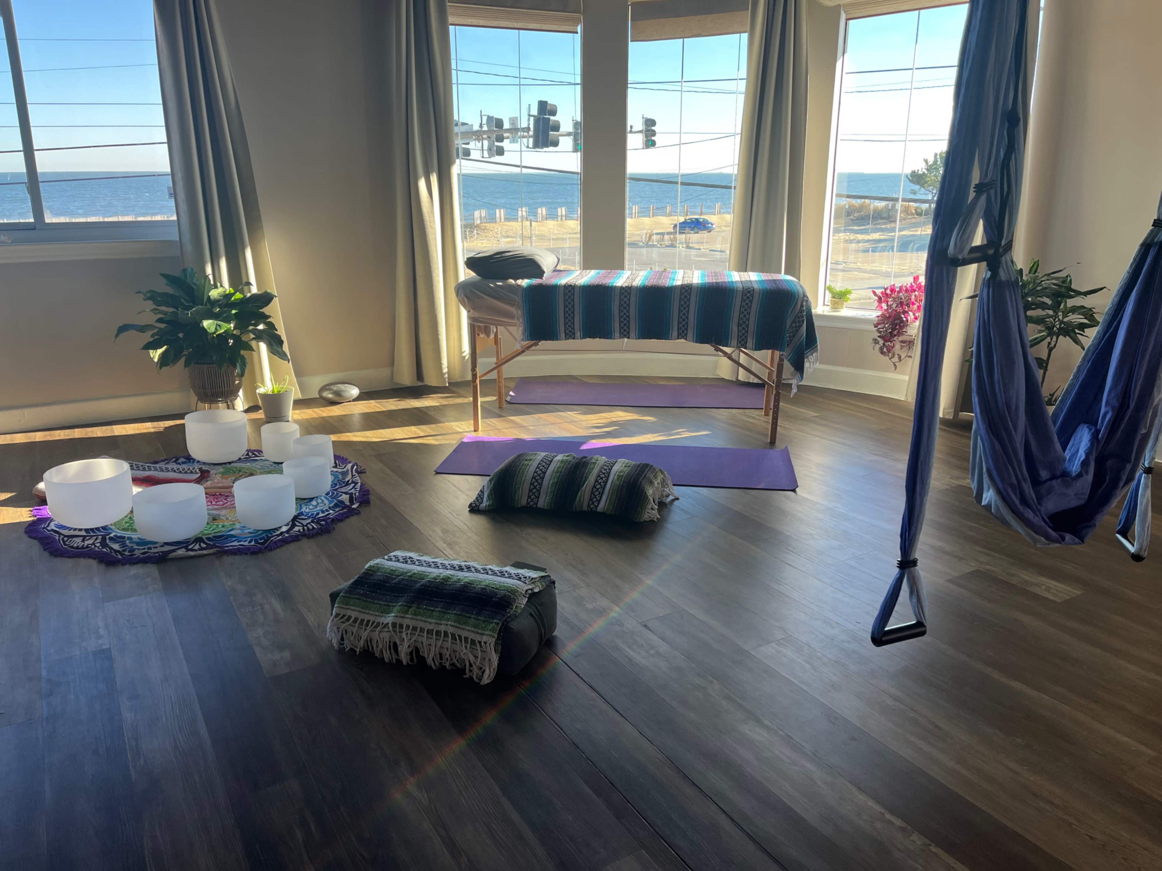 The image shows a serene wellness studio with large windows overlooking the ocean, featuring yoga mats, crystal bowls, and a massage table.