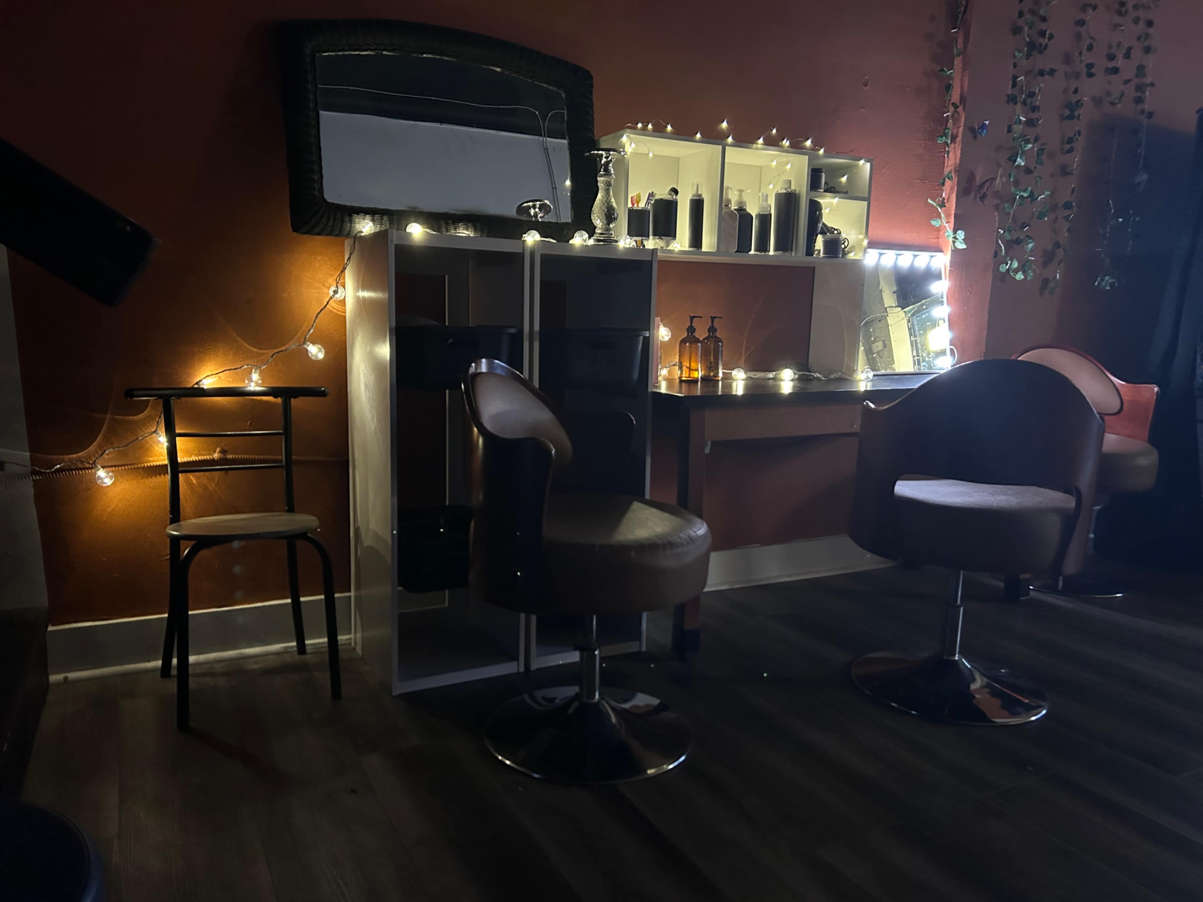 The image features a dimly lit hair salon with two brown salon chairs, a mirrored counter, and decorative string lights against a red wall.
