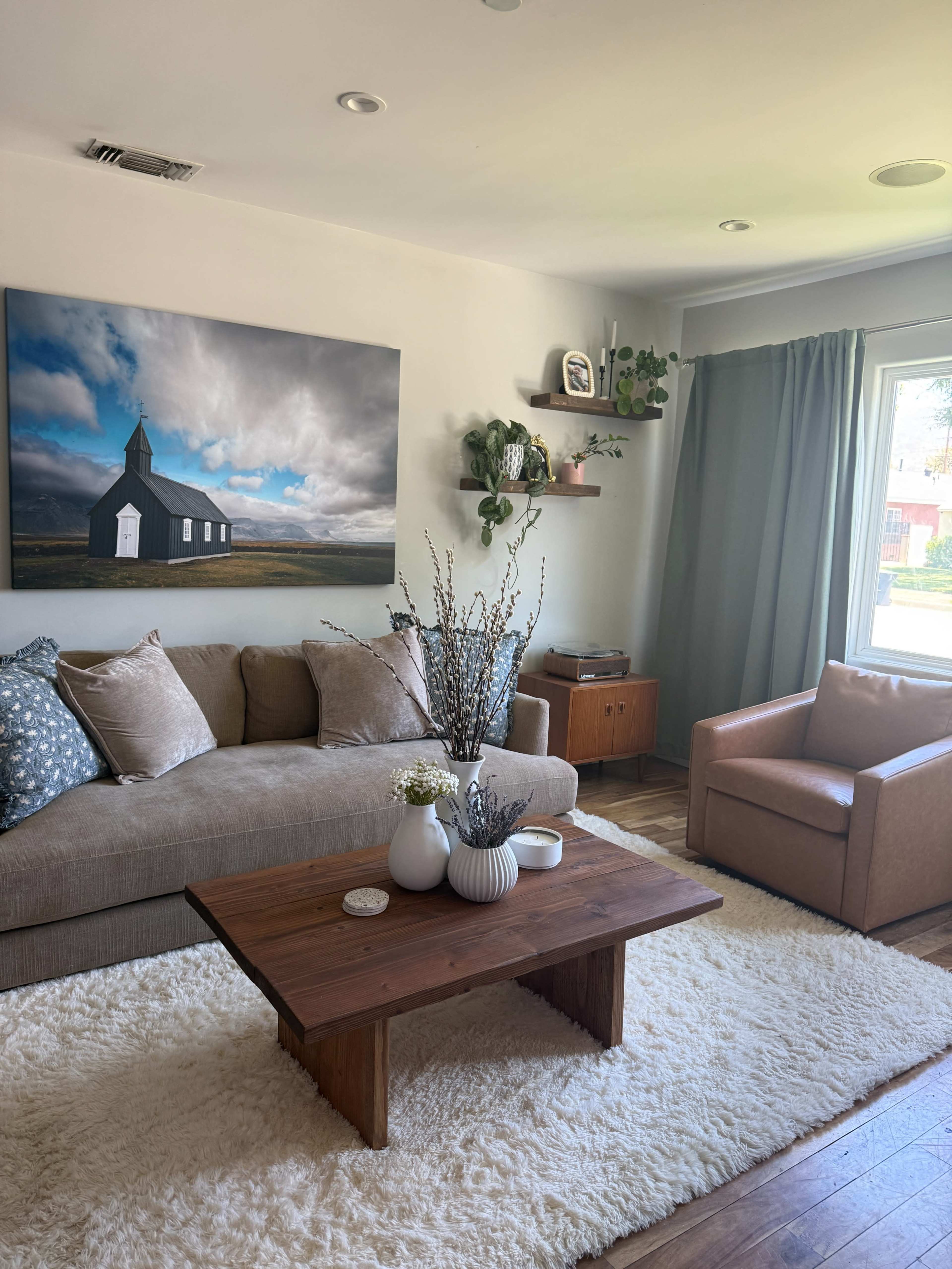 The image shows a cozy living room featuring a beige sofa with decorative pillows, a wooden coffee table, a brown armchair, and a wall-mounted shelf, all arranged on a soft rug under a large photograph of a building.