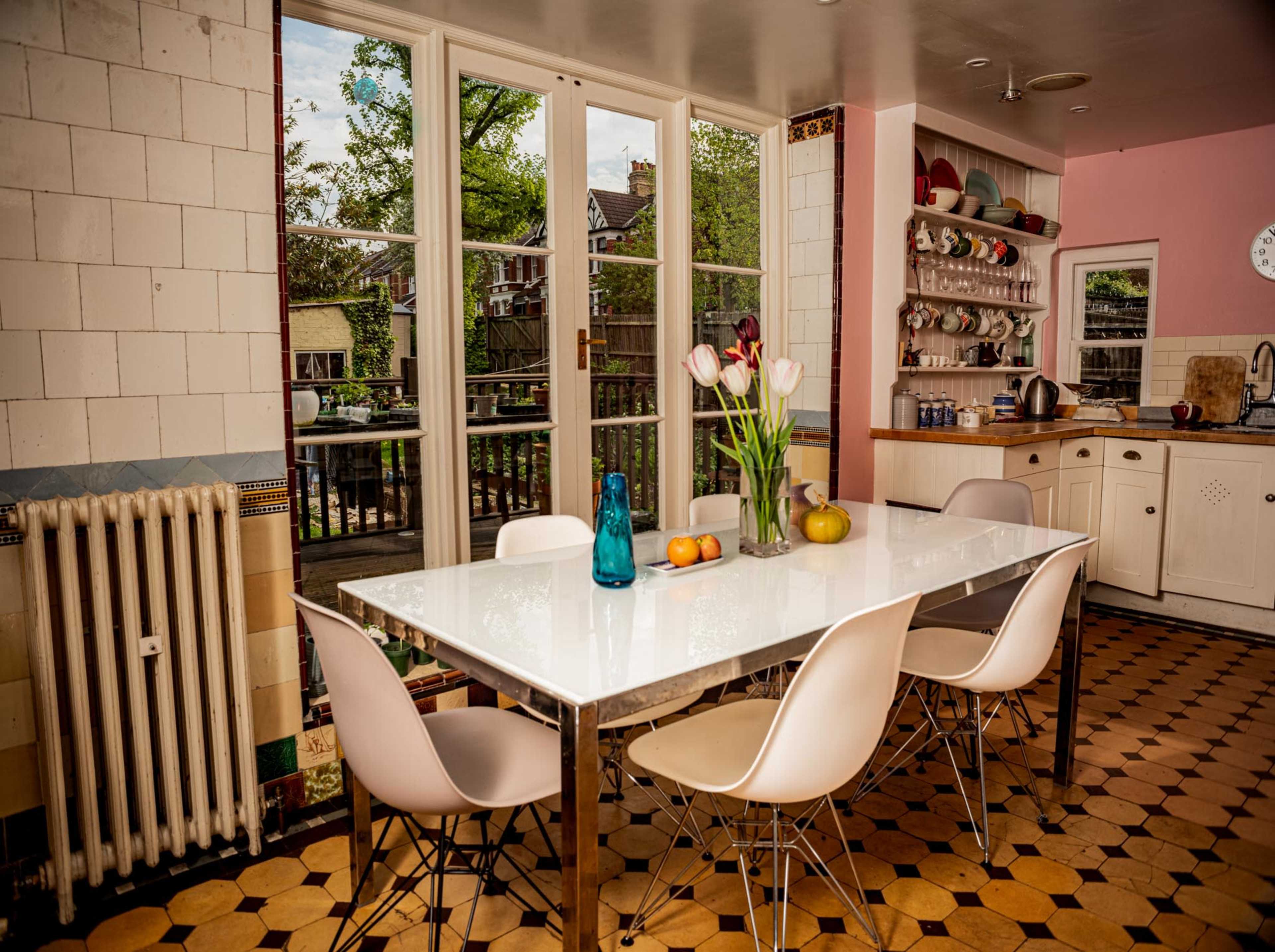 A bright kitchen features a white dining table surrounded by six chairs, with a vase of tulips and fruits on the table, and large windows overlooking a garden.