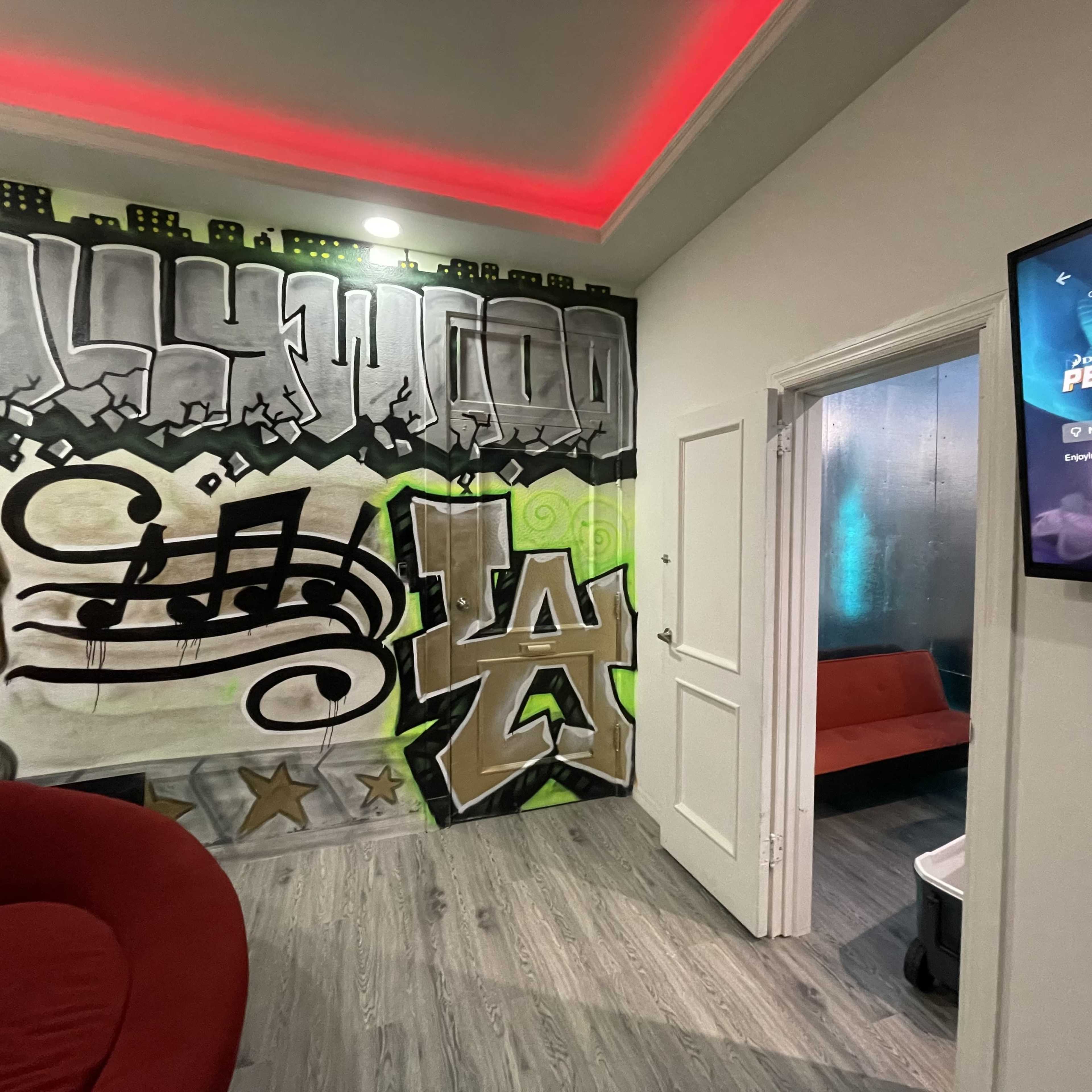 A room featuring graffiti-style wall art with musical notes and "LA" on one wall, while a door leads to another space on the right, and a television is mounted on the opposite wall.
