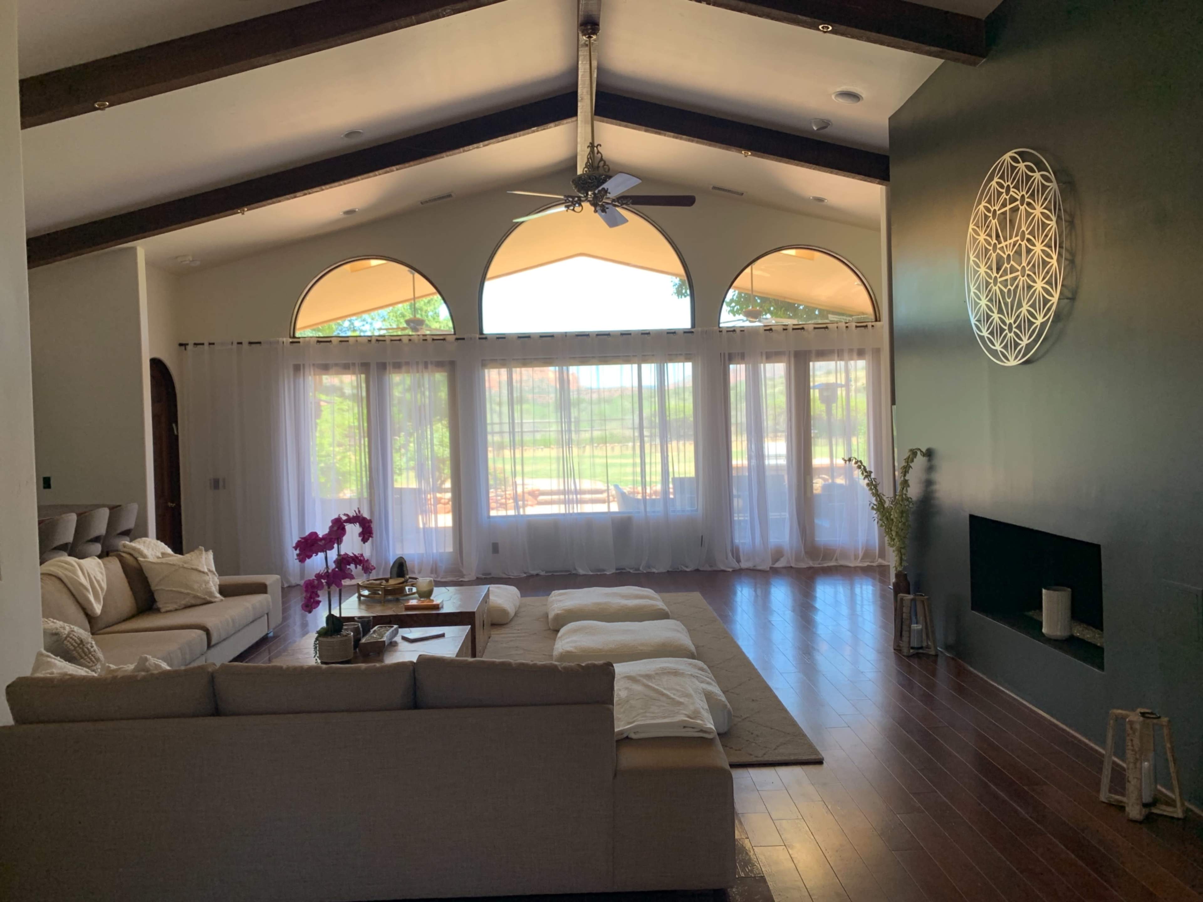 A spacious living room with large windows, a sectional sofa, and a modern wall art piece, featuring wooden beams in the ceiling.