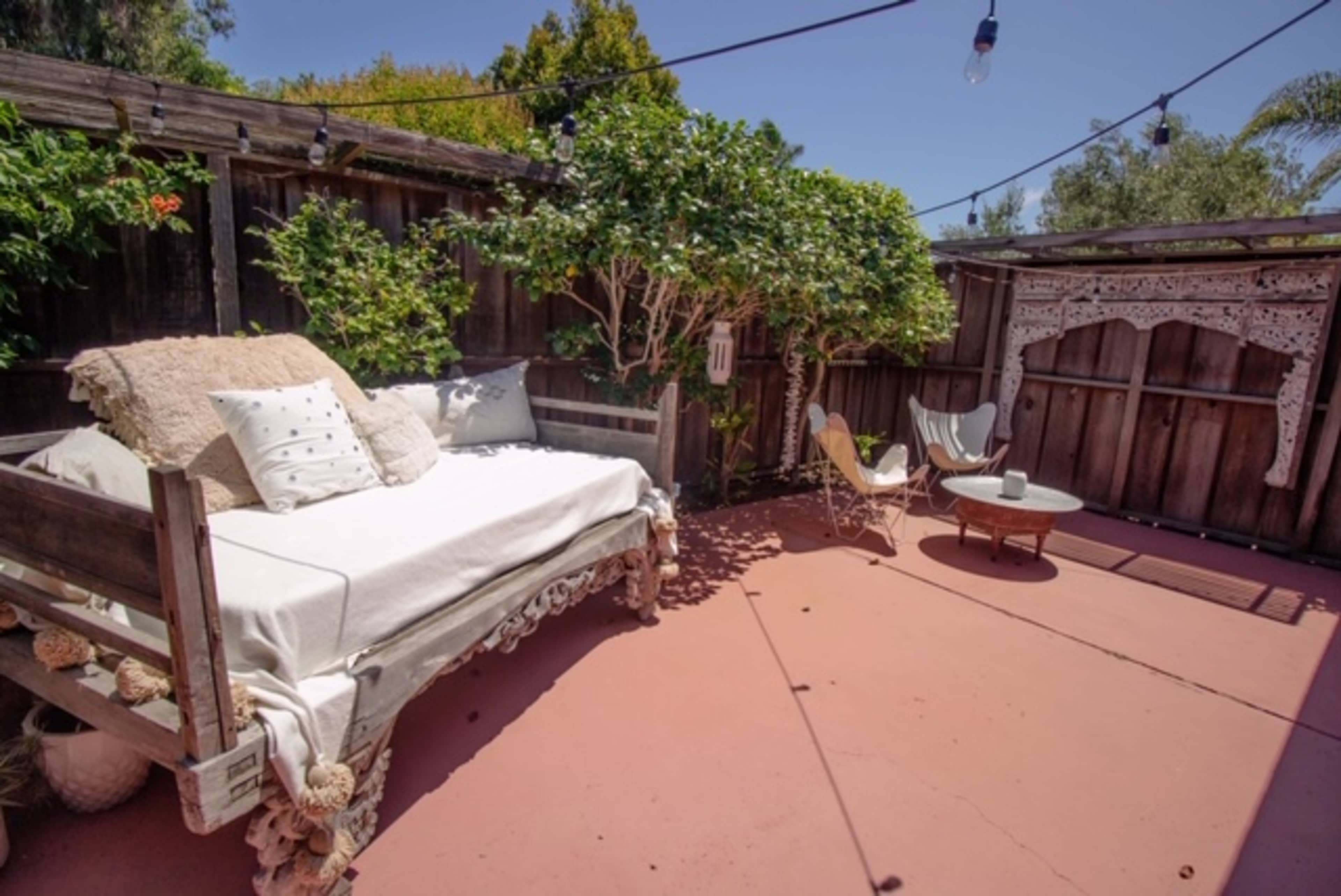 A patio features a daybed with decorative pillows, a small table, and a chair surrounded by greenery and wooden fences.