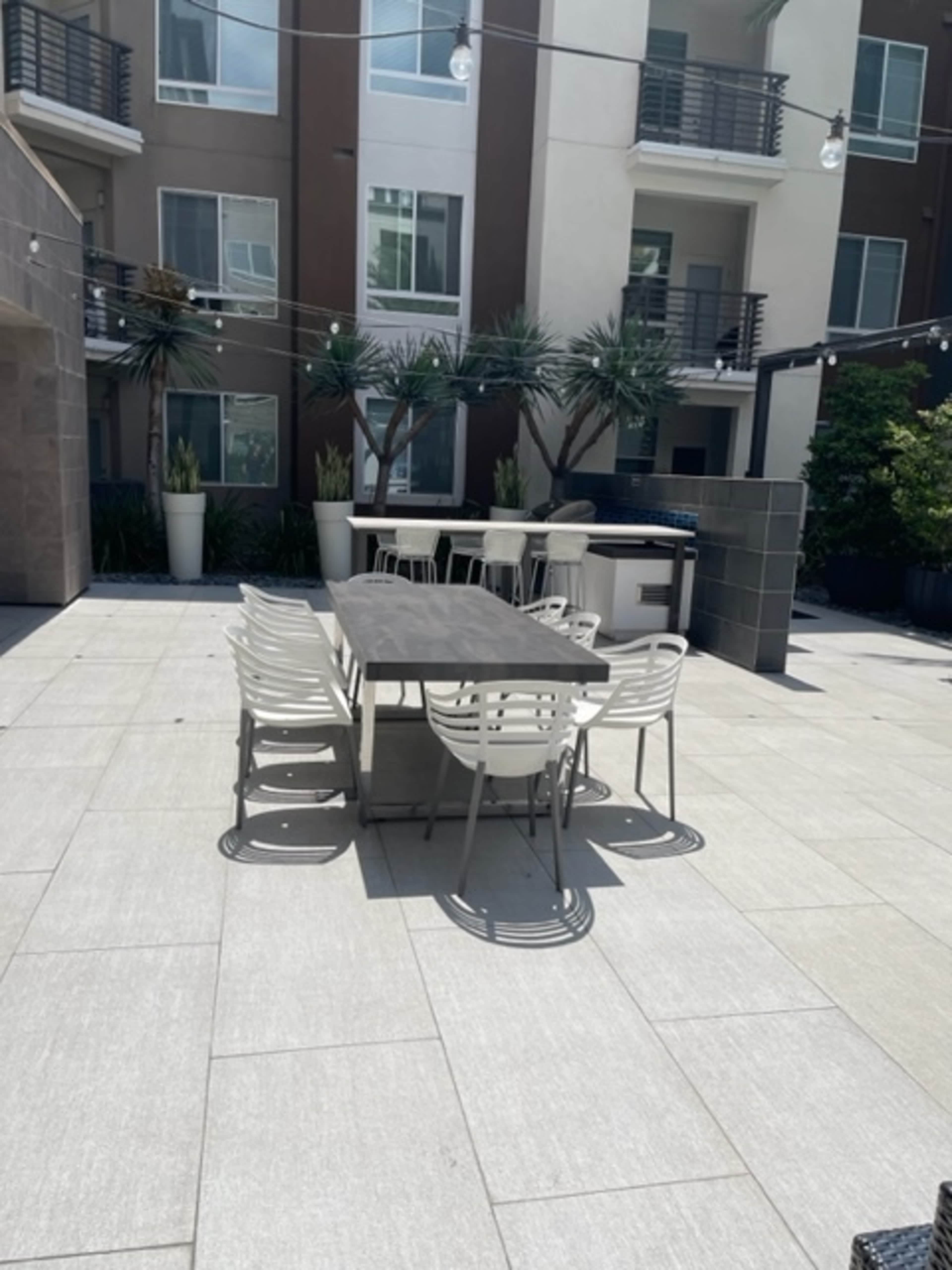 A dining table with chairs is positioned on a tiled patio surrounded by modern apartment buildings and planters.
