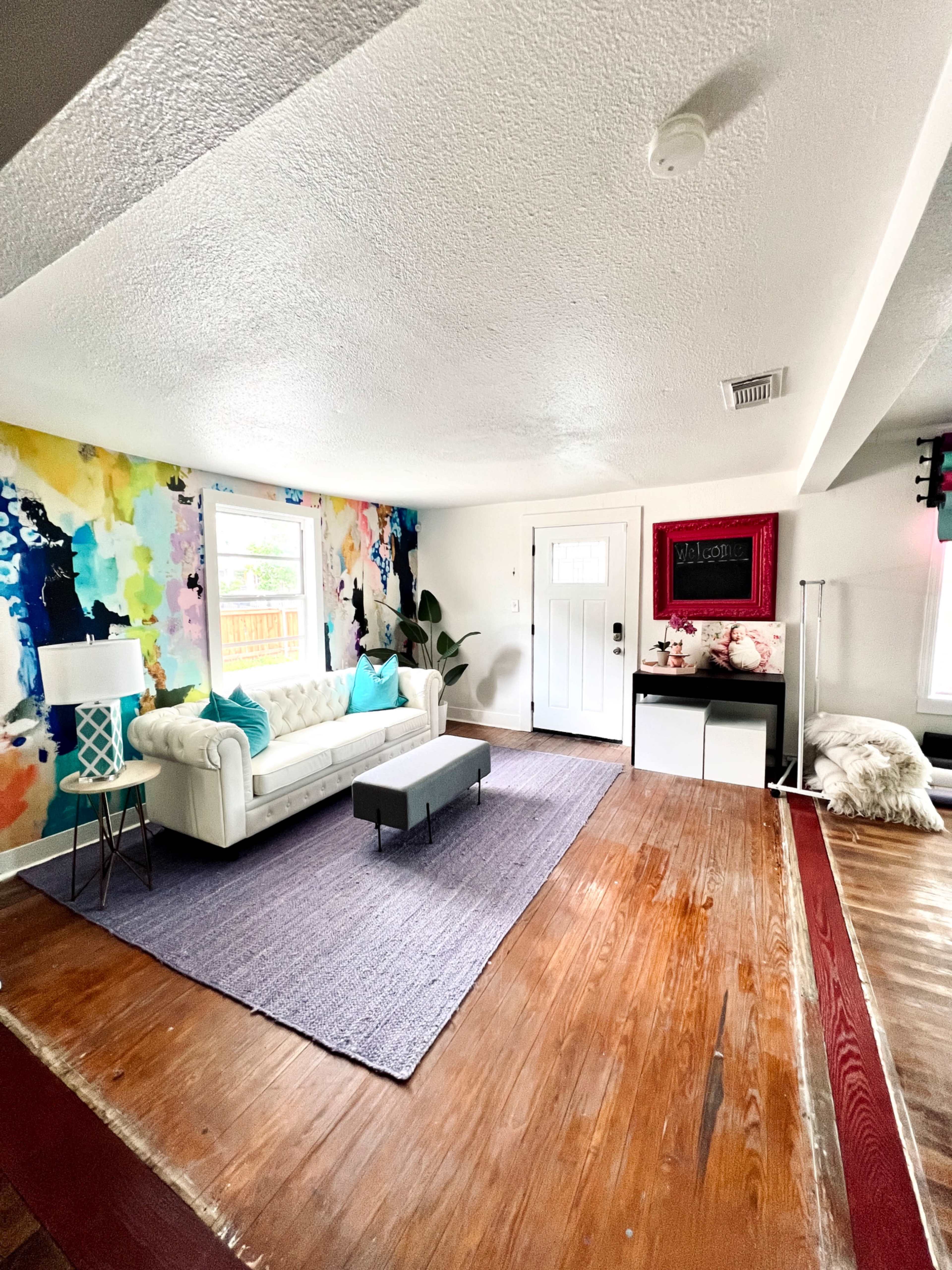 The image shows a spacious living room with a white tufted sofa, colorful abstract wall art, a brown wooden floor, and a small gray coffee table in the center.