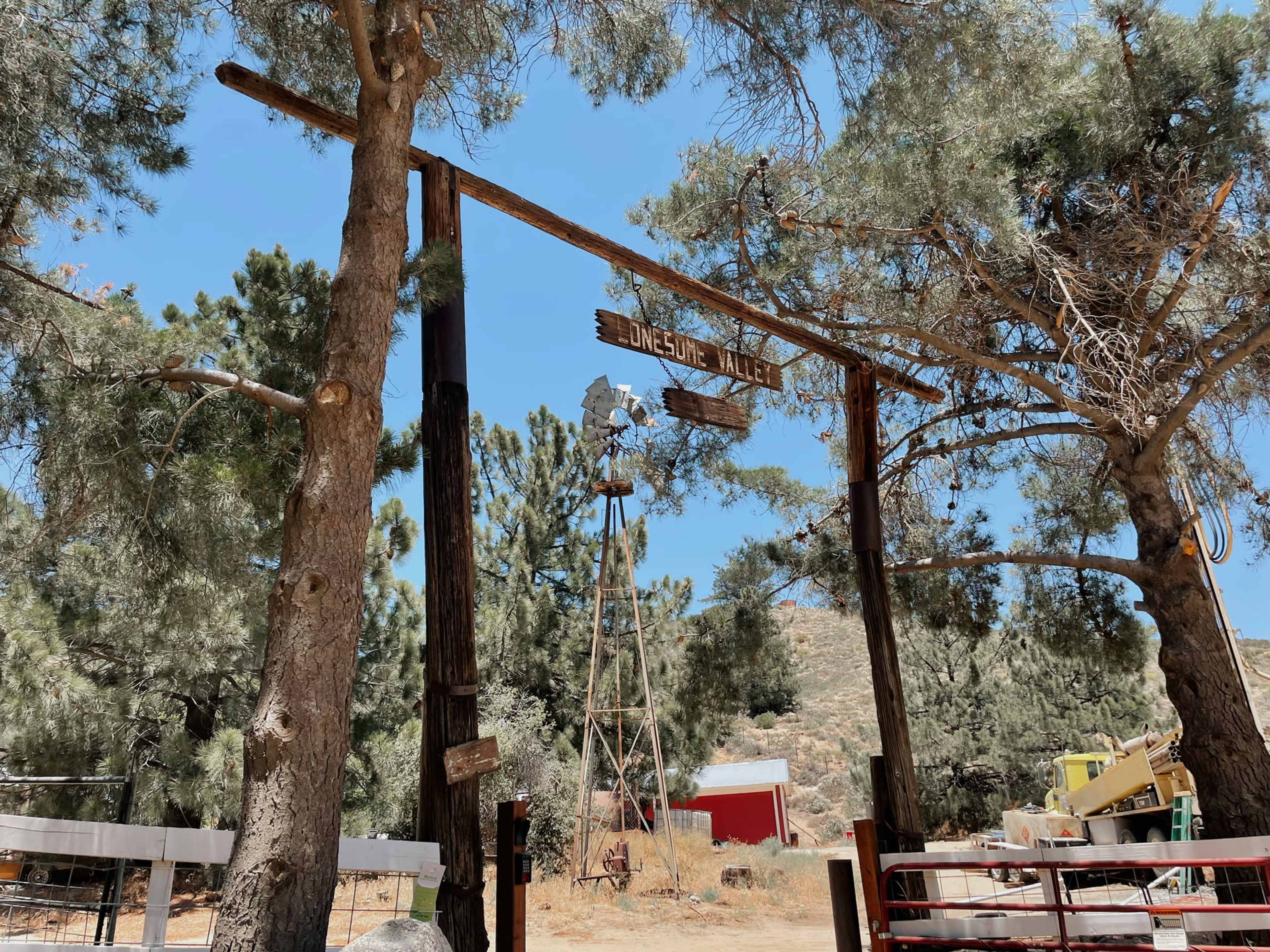A wooden archway labeled "Lonesome Trail" stands among tall pine trees, with a windmill and a red building visible in the background.