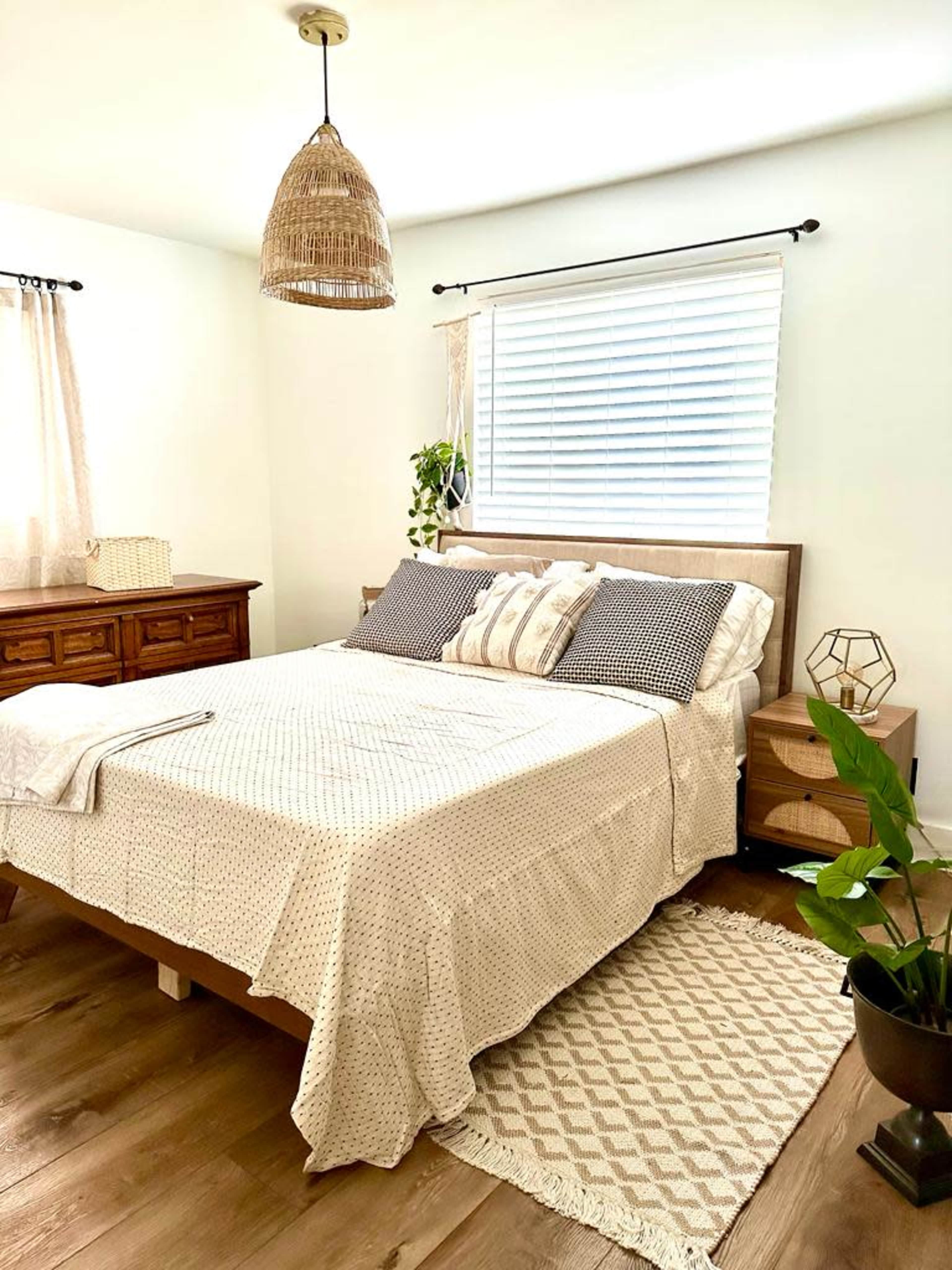 The image shows a bright bedroom featuring a bed with patterned bedding, wooden furniture, and a woven pendant light.