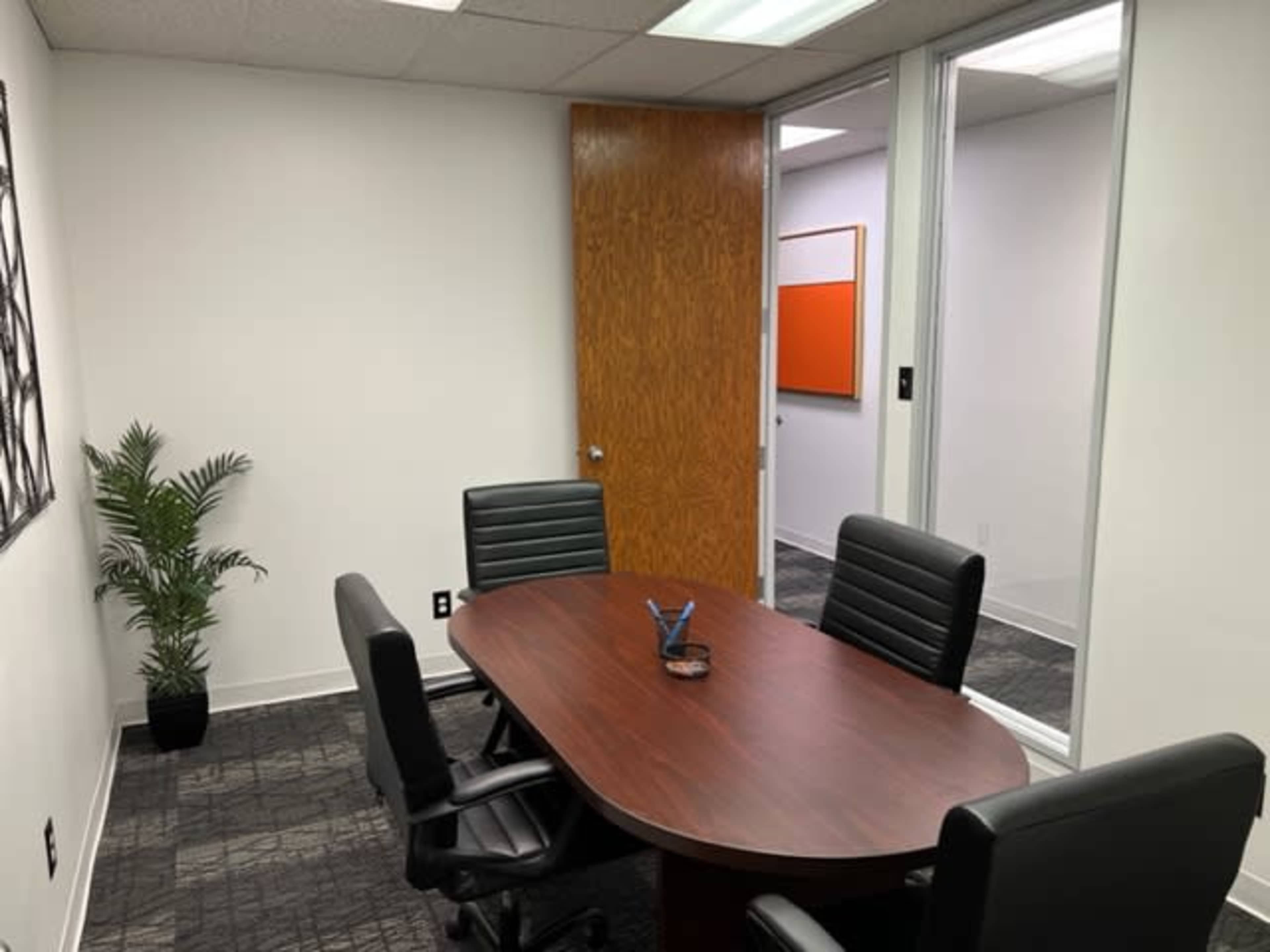 A small conference room with a wooden oval table and four black chairs, accompanied by a potted plant in one corner and a door leading to an adjacent room.