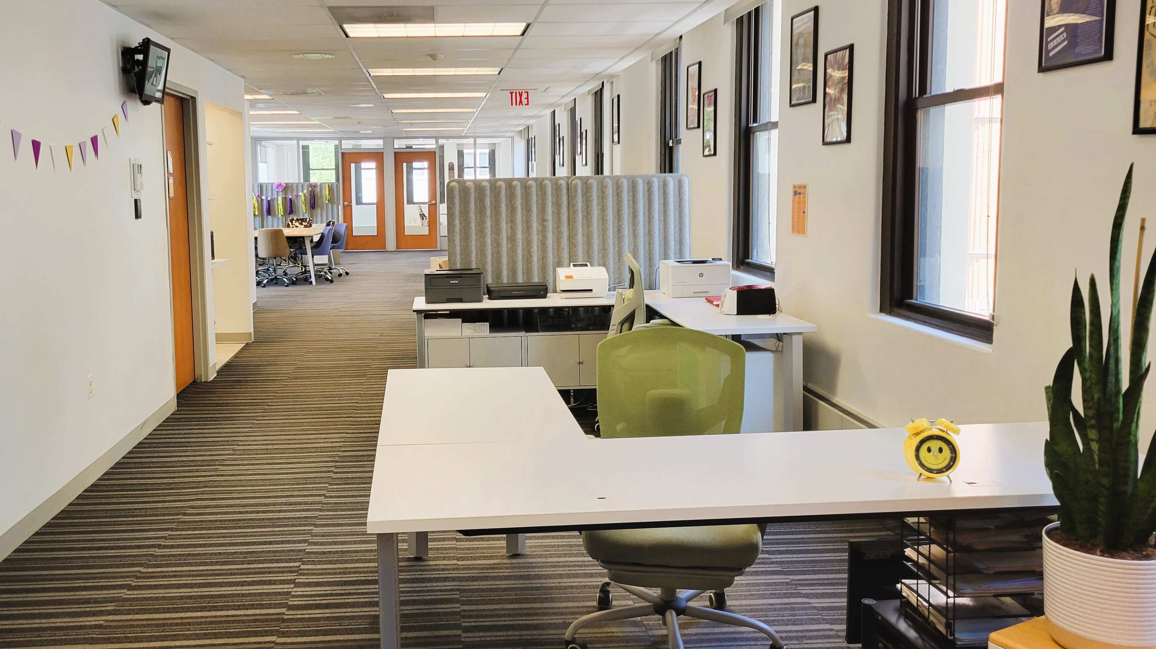 A bright office corridor with workstations, including desks and printers, lined on either side and framed artwork on the walls.