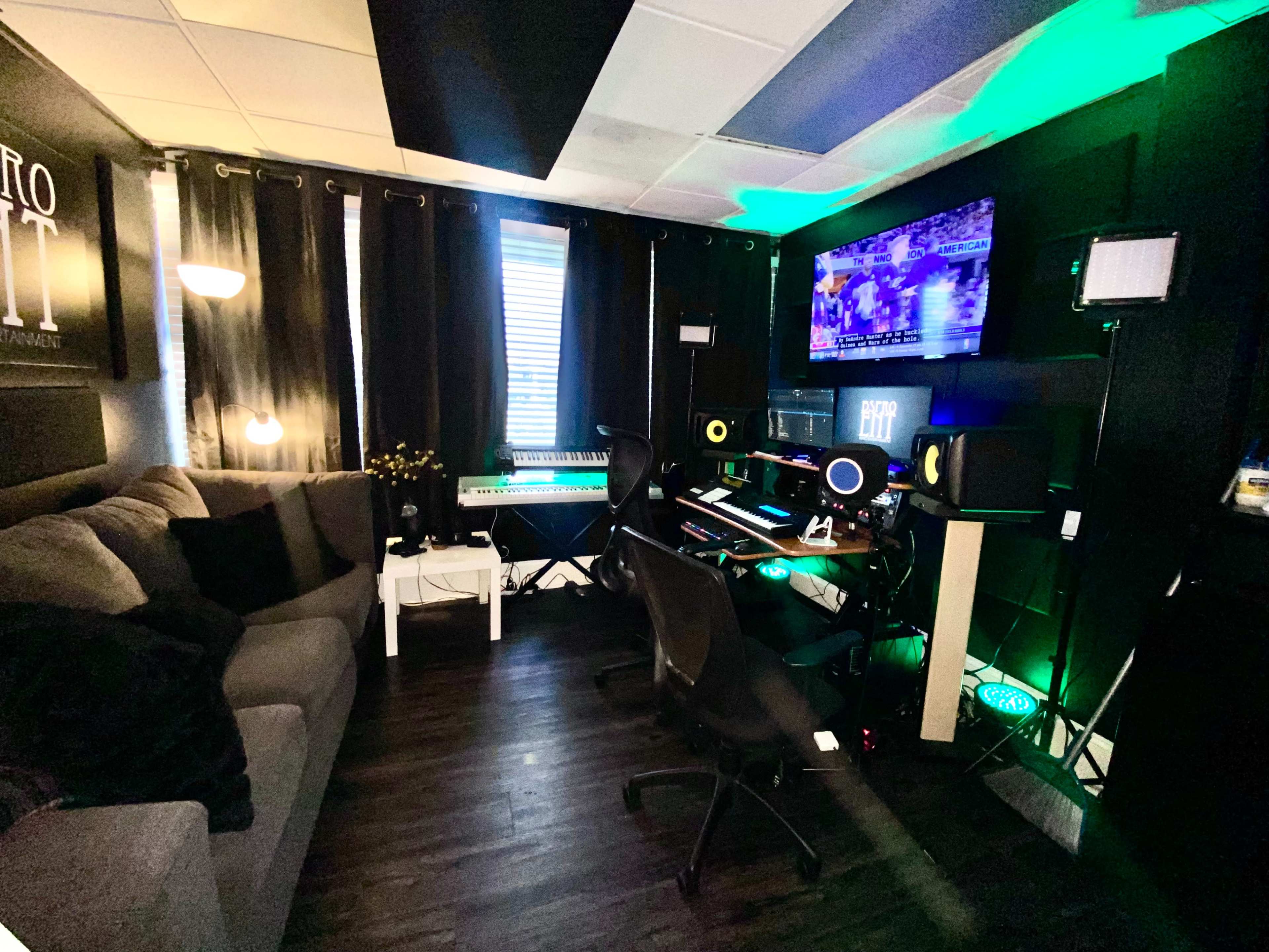 The image shows a small music studio featuring a couch, a keyboard, and a workstation with multiple monitors and speakers, illuminated by green LED lights.