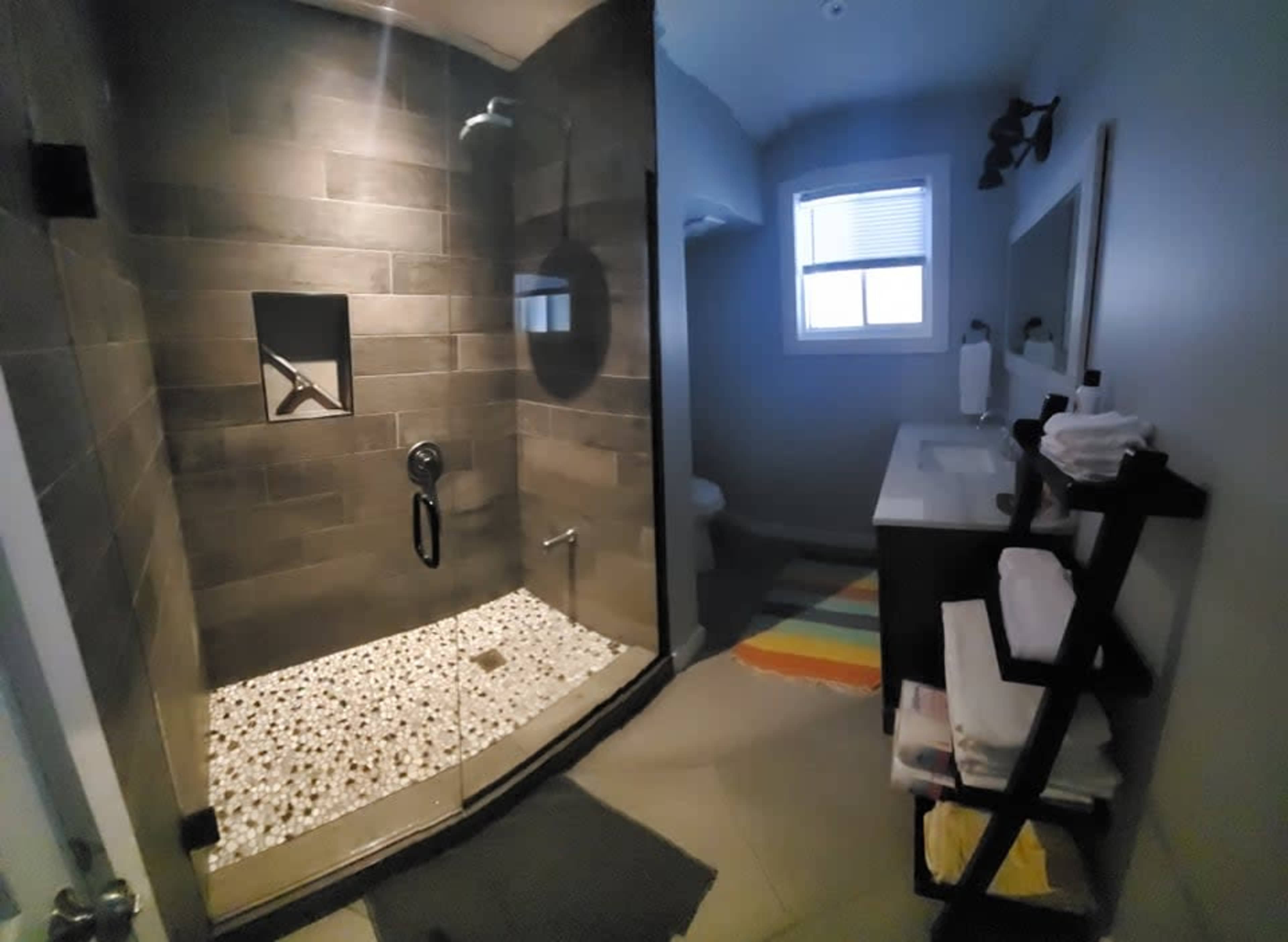 A modern bathroom with a glass shower enclosure featuring stone tile and a pebble-style floor, alongside a vanity area and a small window.