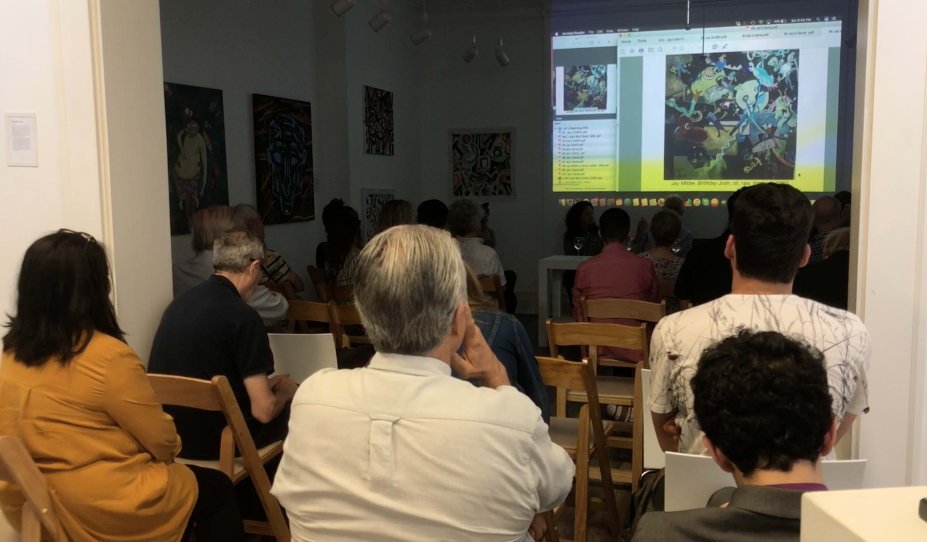 A group of people sits in a room facing a large screen displaying colorful artwork.