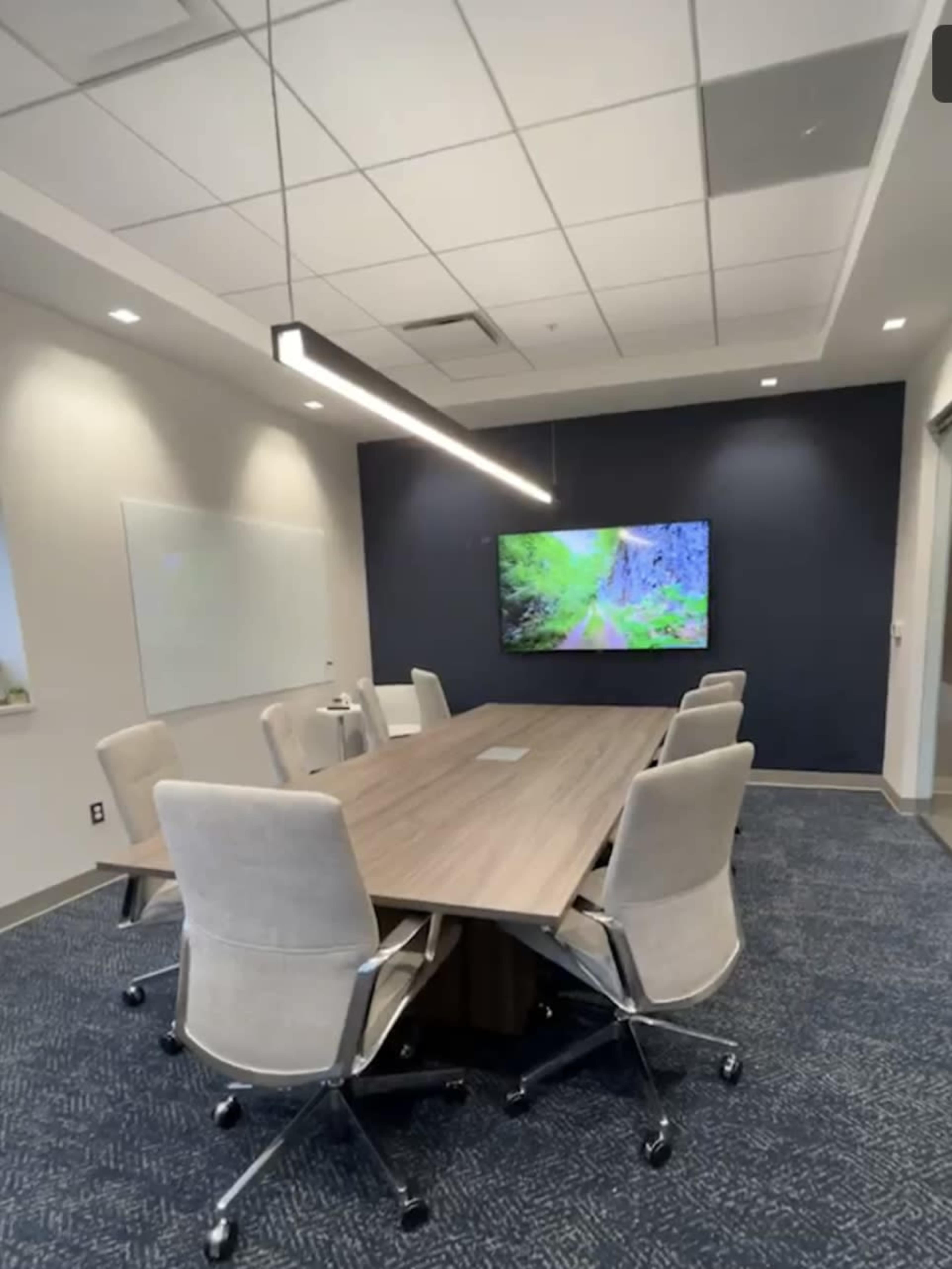 A modern conference room featuring a long table surrounded by ergonomic chairs and a large screen displaying a nature scene on the wall.