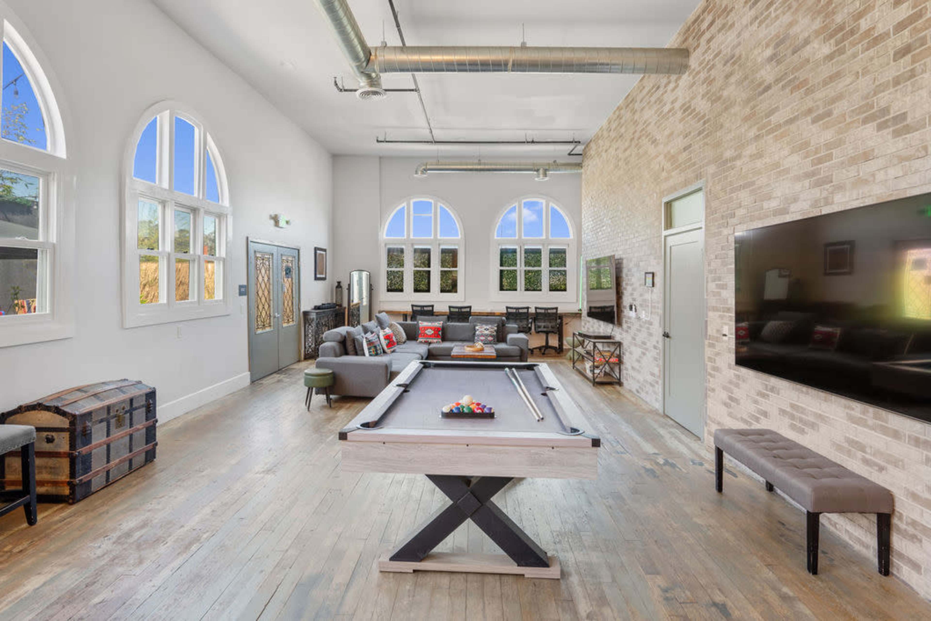 The image shows a modern game room featuring a pool table in the center, with a seating area and large windows that allow natural light to enter.