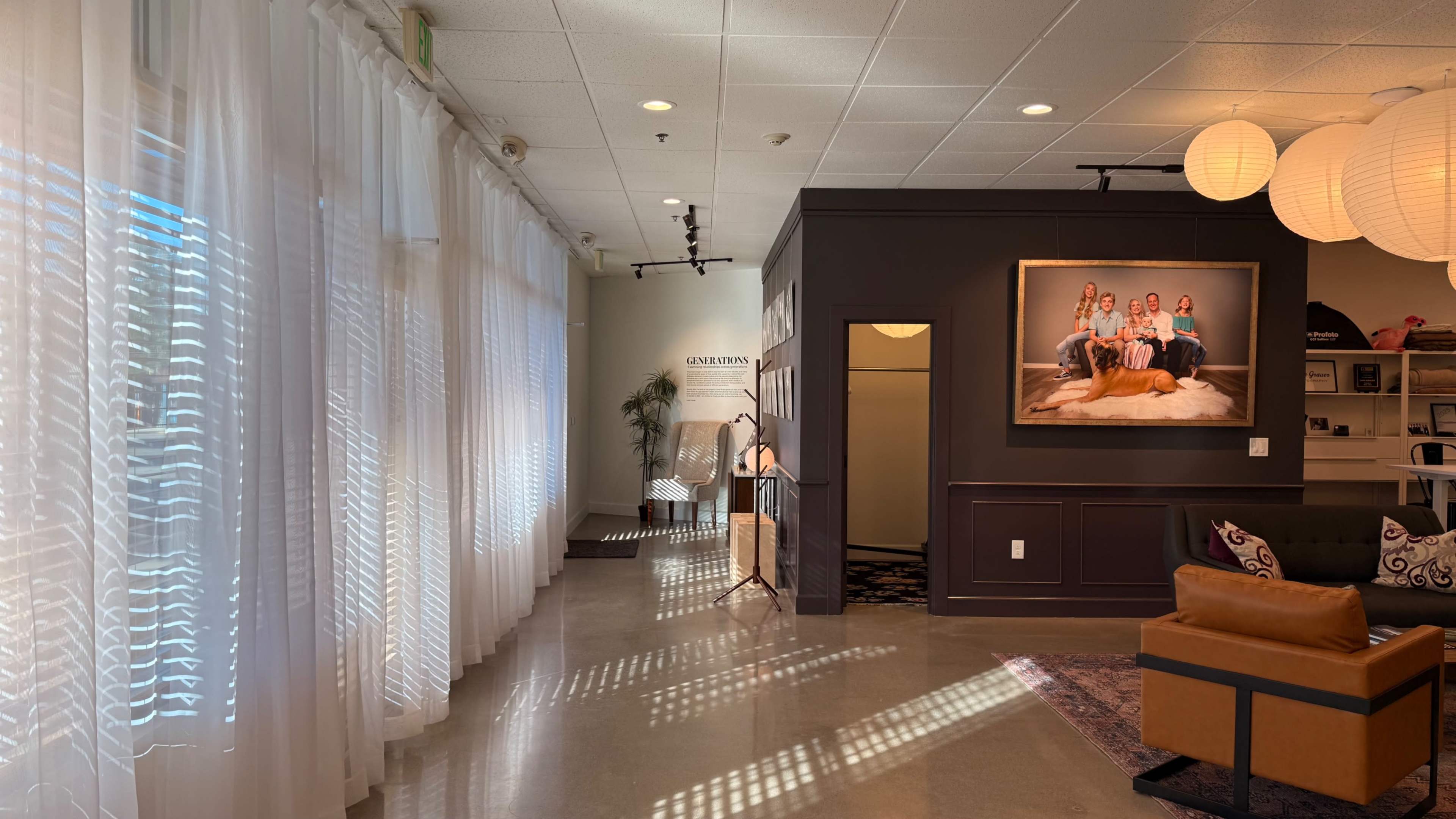 The image shows a spacious indoor area with large windows dressed in sheer curtains, a dark accent wall featuring a group portrait, and a cozy seating arrangement.