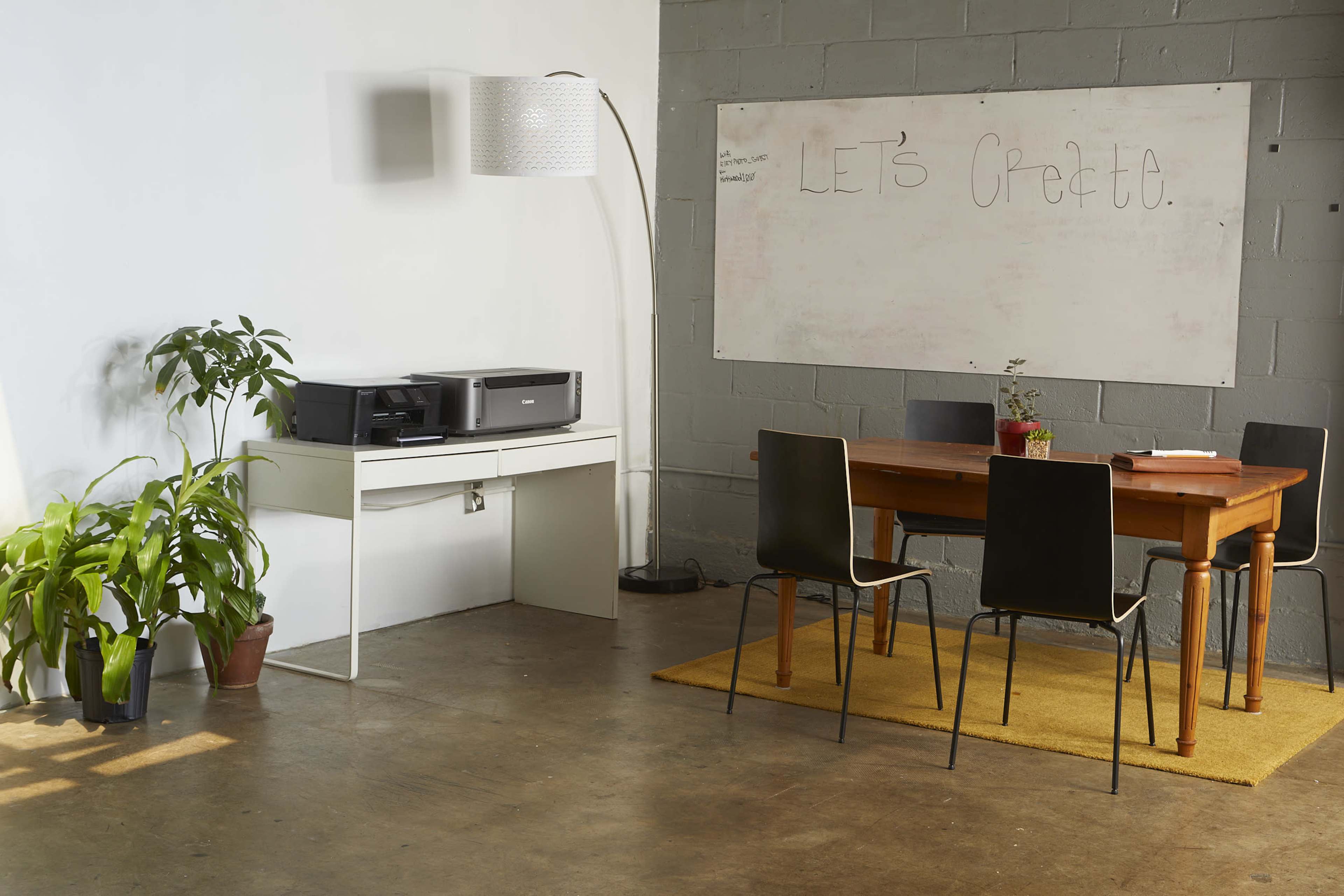 A minimalistic office space features a wooden table with four black chairs, a printer on a white desk, a plant in a pot, and a whiteboard with the phrase "LET'S Create."
