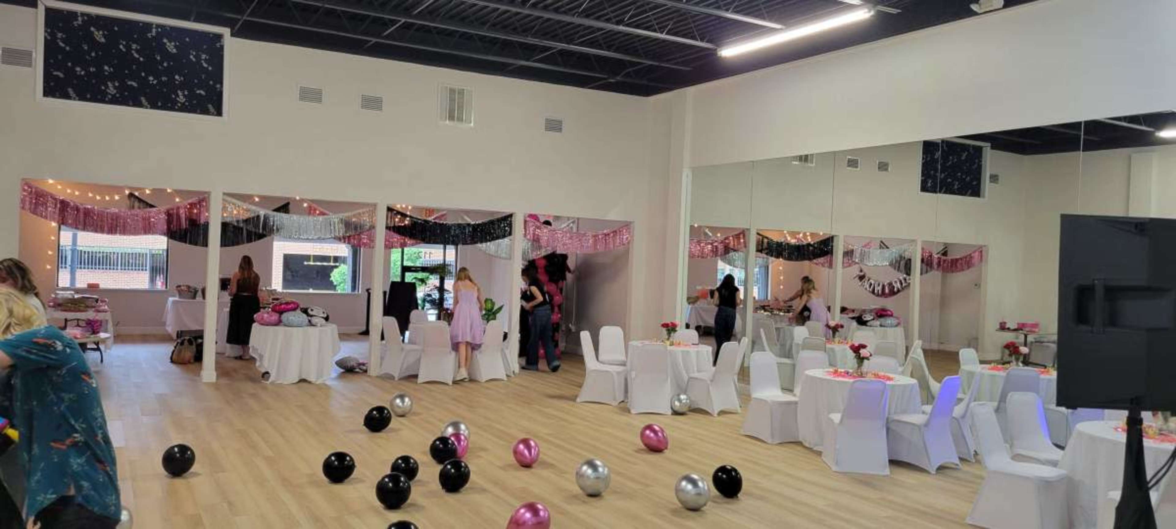 The image shows a decorated venue with tables and chairs set up for an event, featuring balloons on the floor and a mirrored wall reflecting the scene.