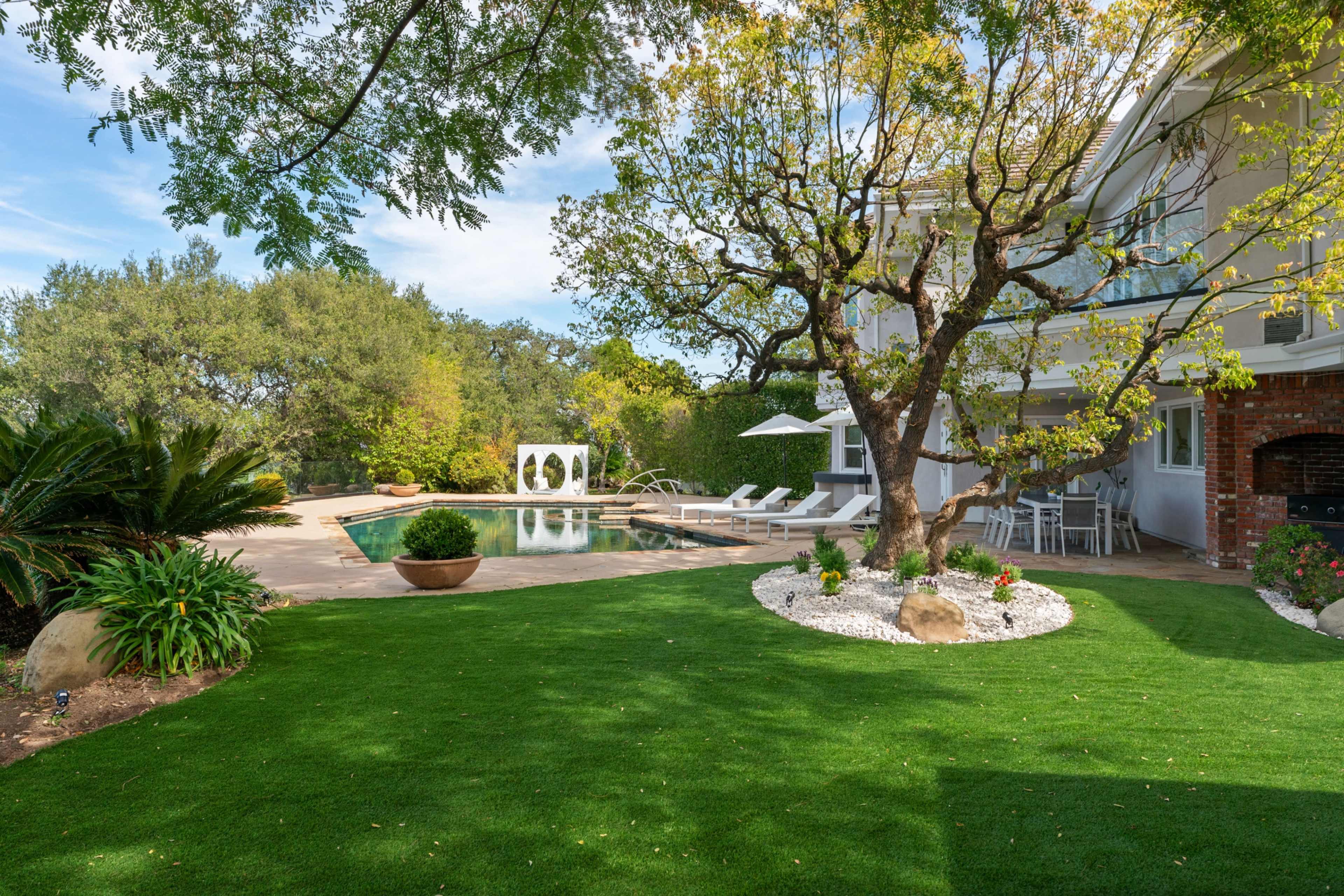 The image shows a landscaped backyard featuring a pool, lounge chairs, a tree, and a seating area surrounded by greenery.