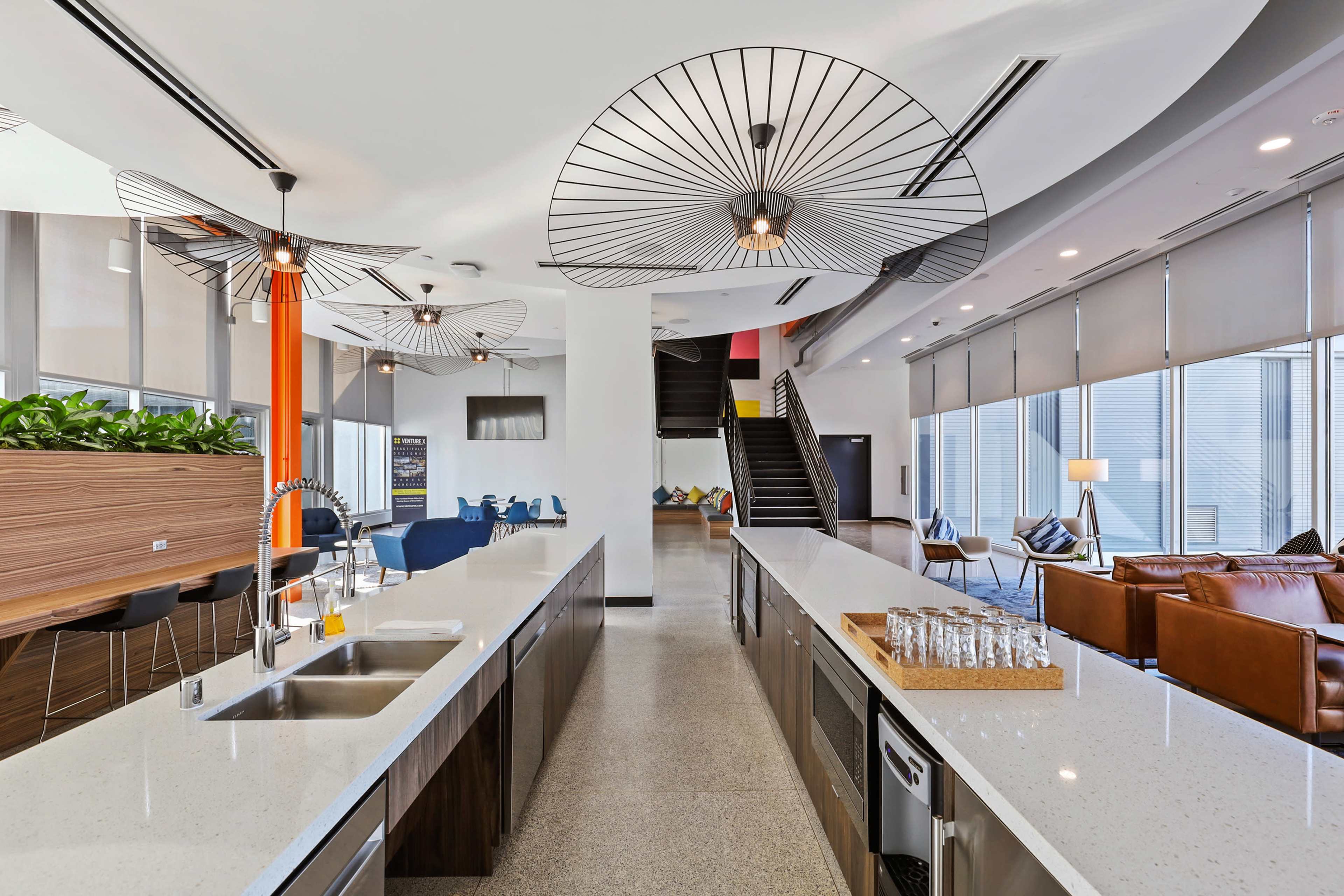 The image shows a modern open space with a kitchen area featuring a long countertop, decorative pendant lights, and seating areas with a staircase in the background.