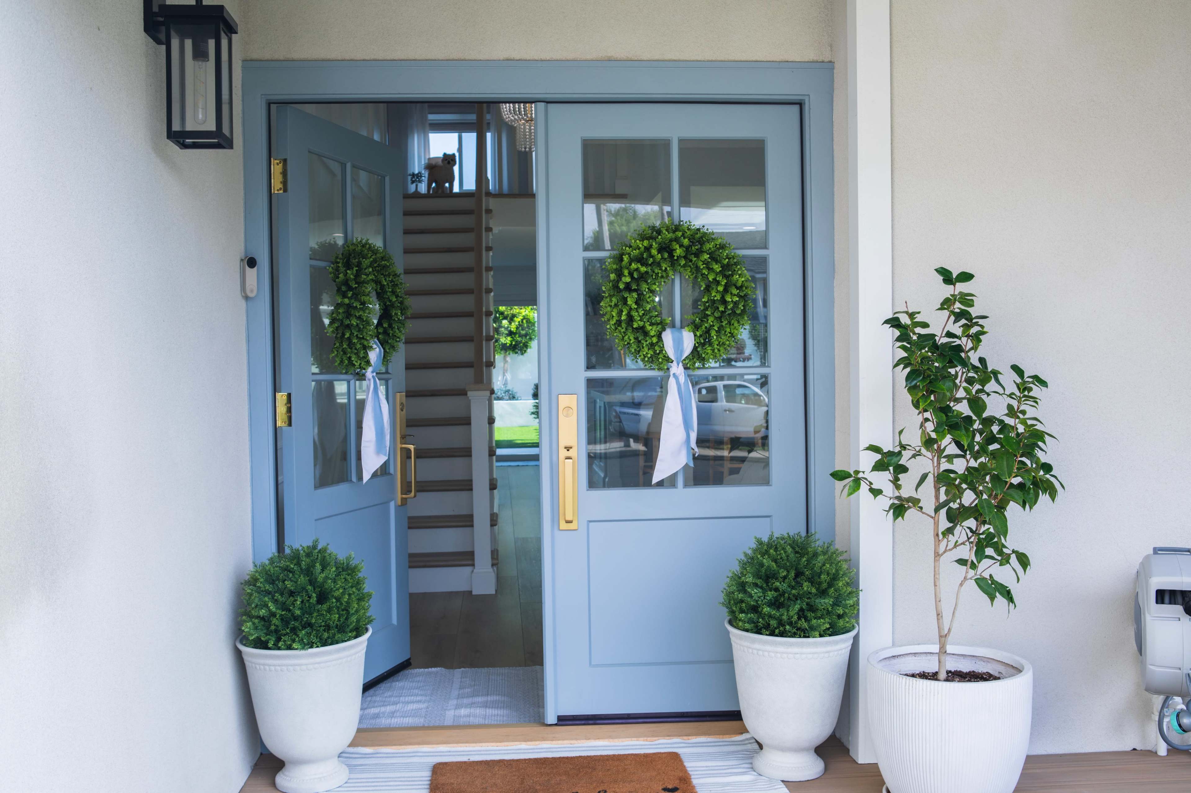The image shows a pair of blue double doors with glass panels, adorned with green wreaths, flanked by potted plants on either side.
