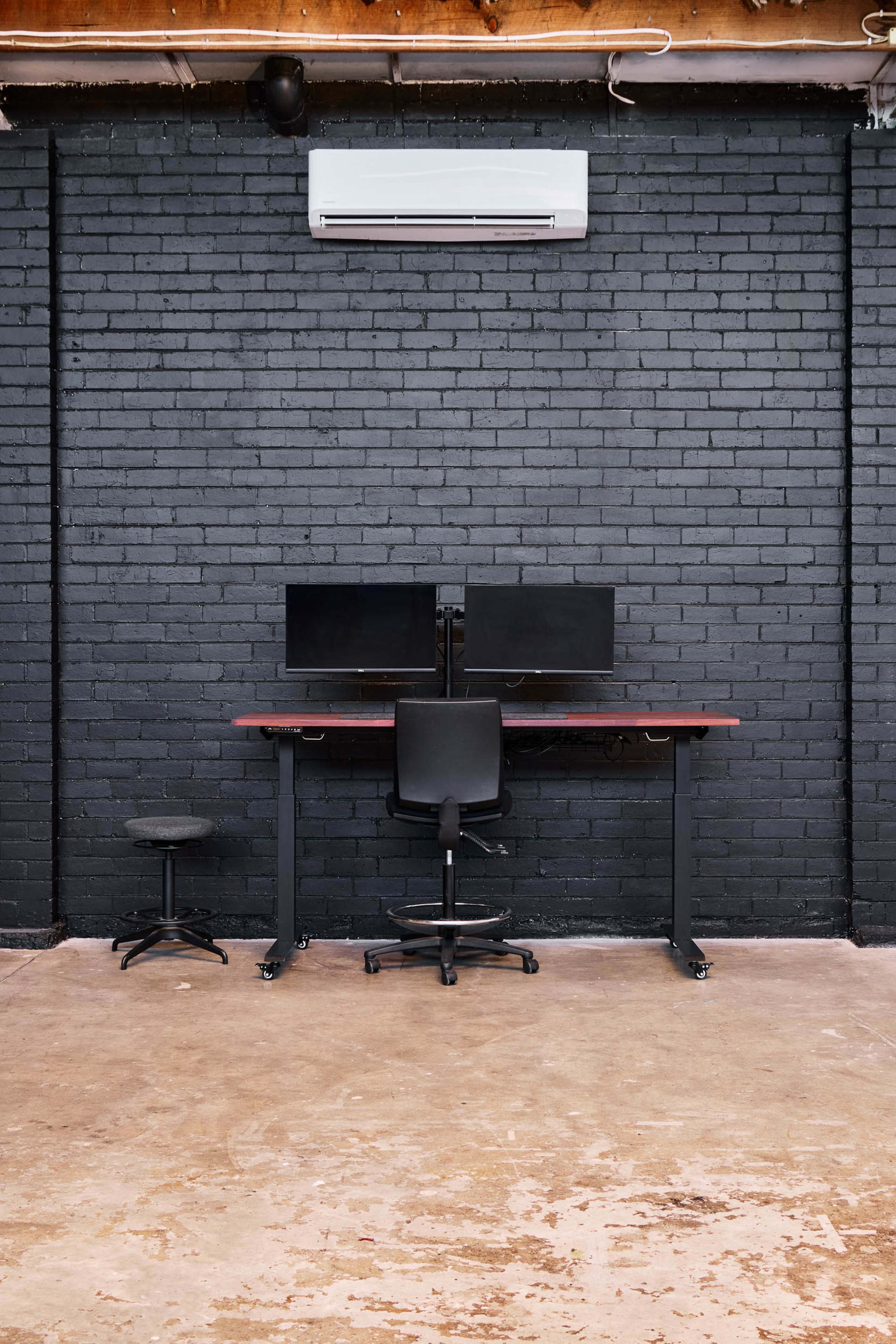 A minimalist workspace features a desk with two monitors and a chair against a black brick wall.