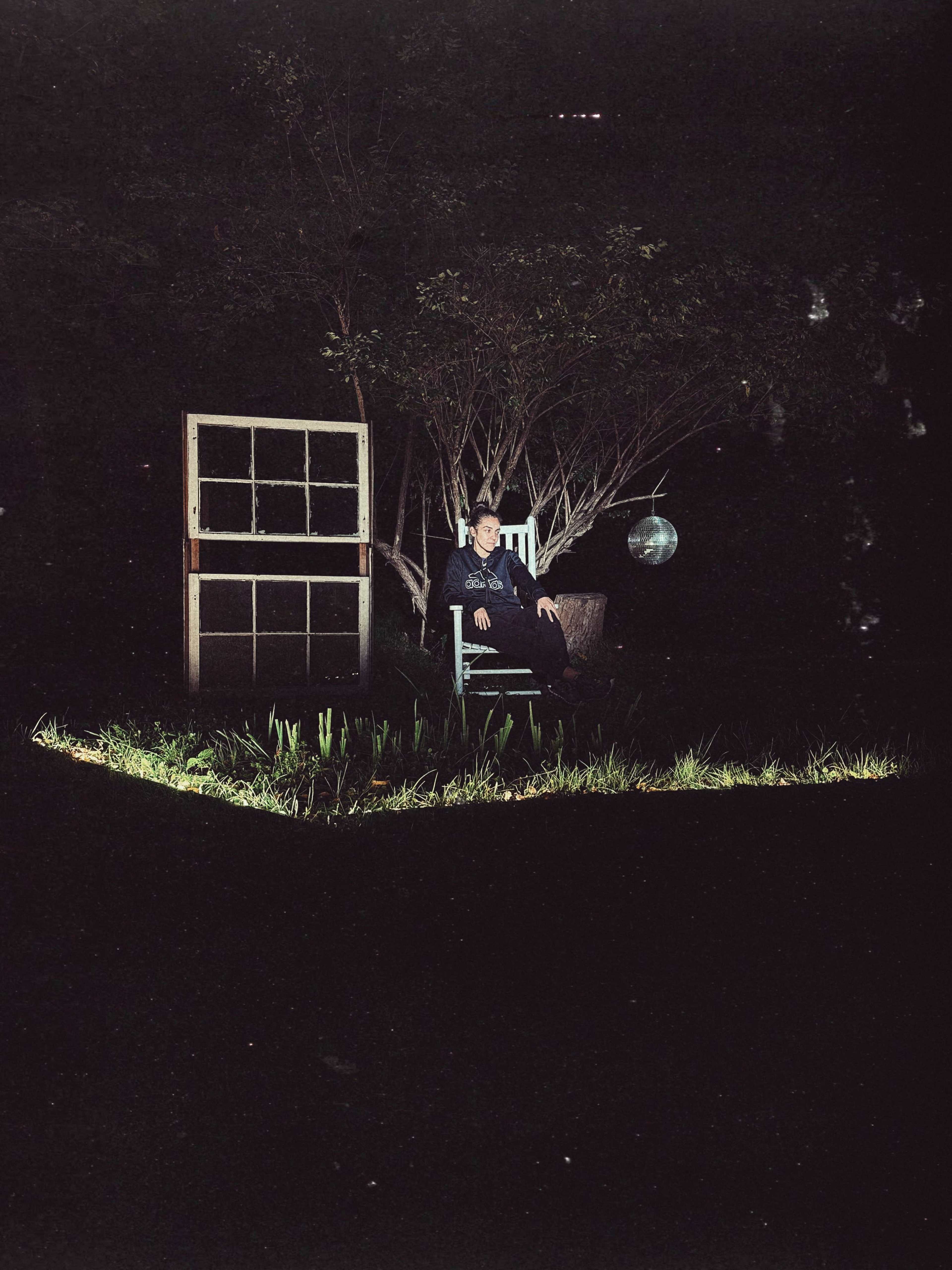 A person sits on a chair in a dark outdoor space, illuminated by light, with a tree and an old window frame in the background.