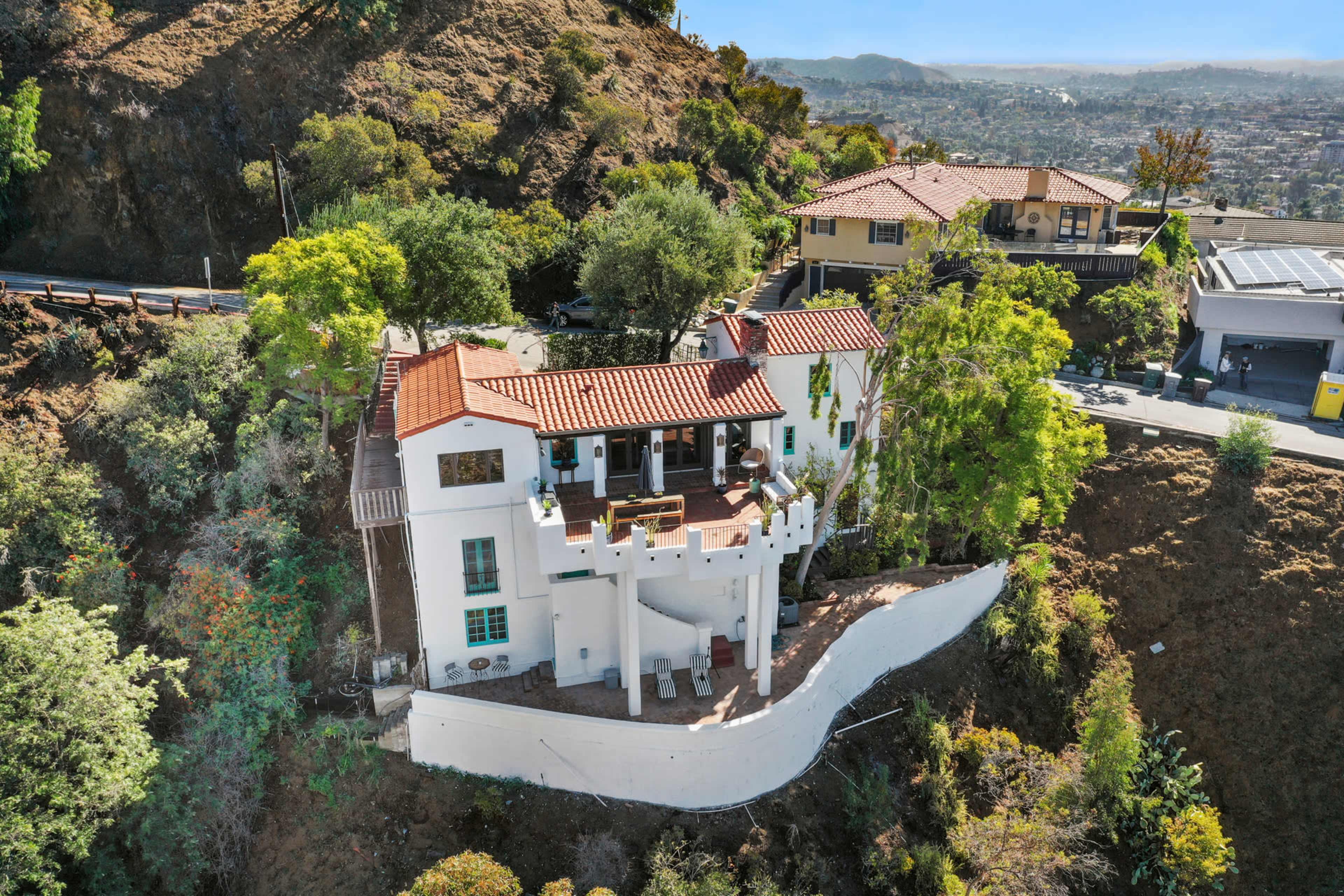 A white stucco house with a red-tiled roof is situated on a hillside surrounded by trees, with a view of the valley below.