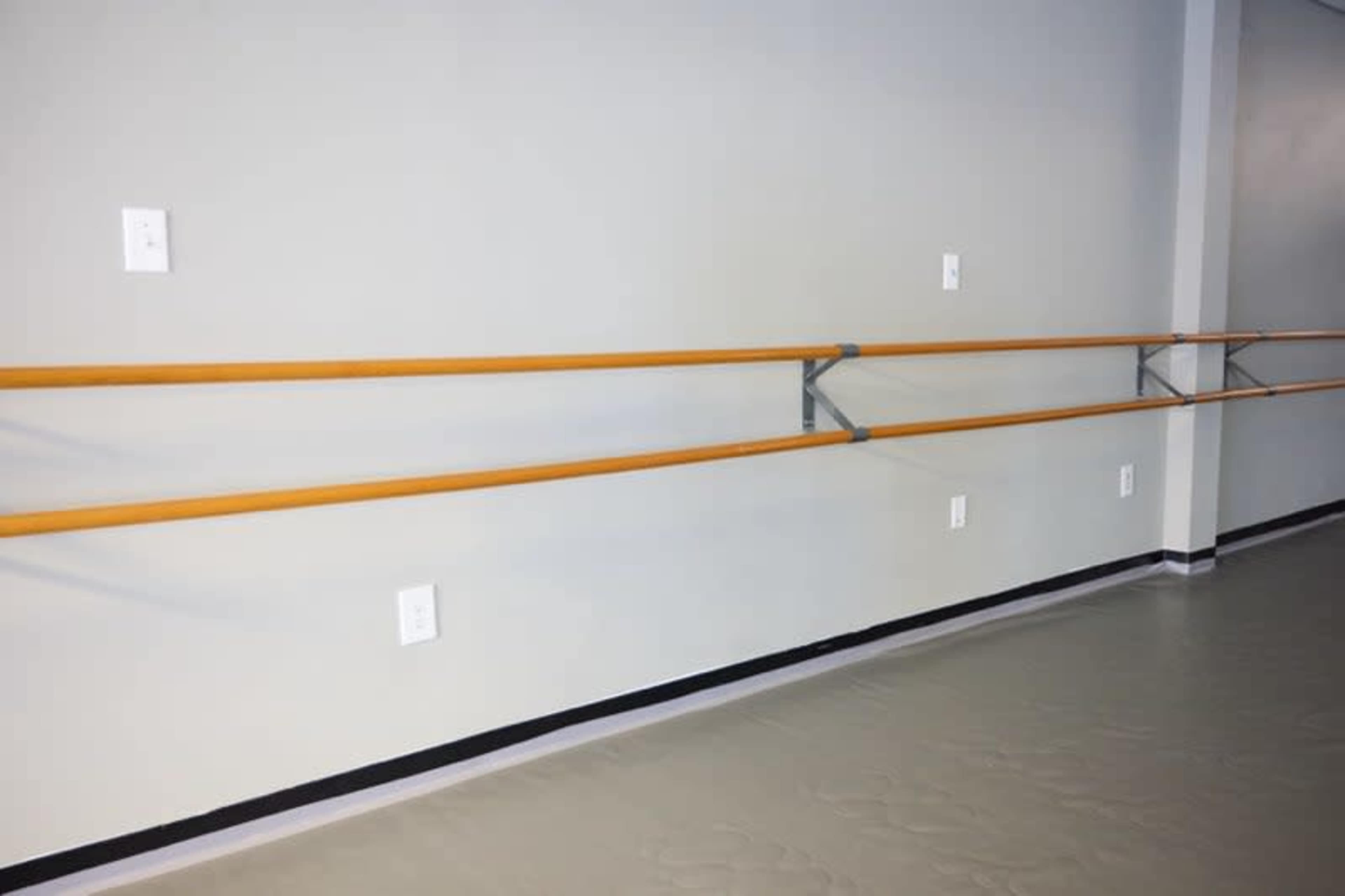 A dance studio interior with a wooden ballet bar mounted on a gray wall and electrical outlets nearby.