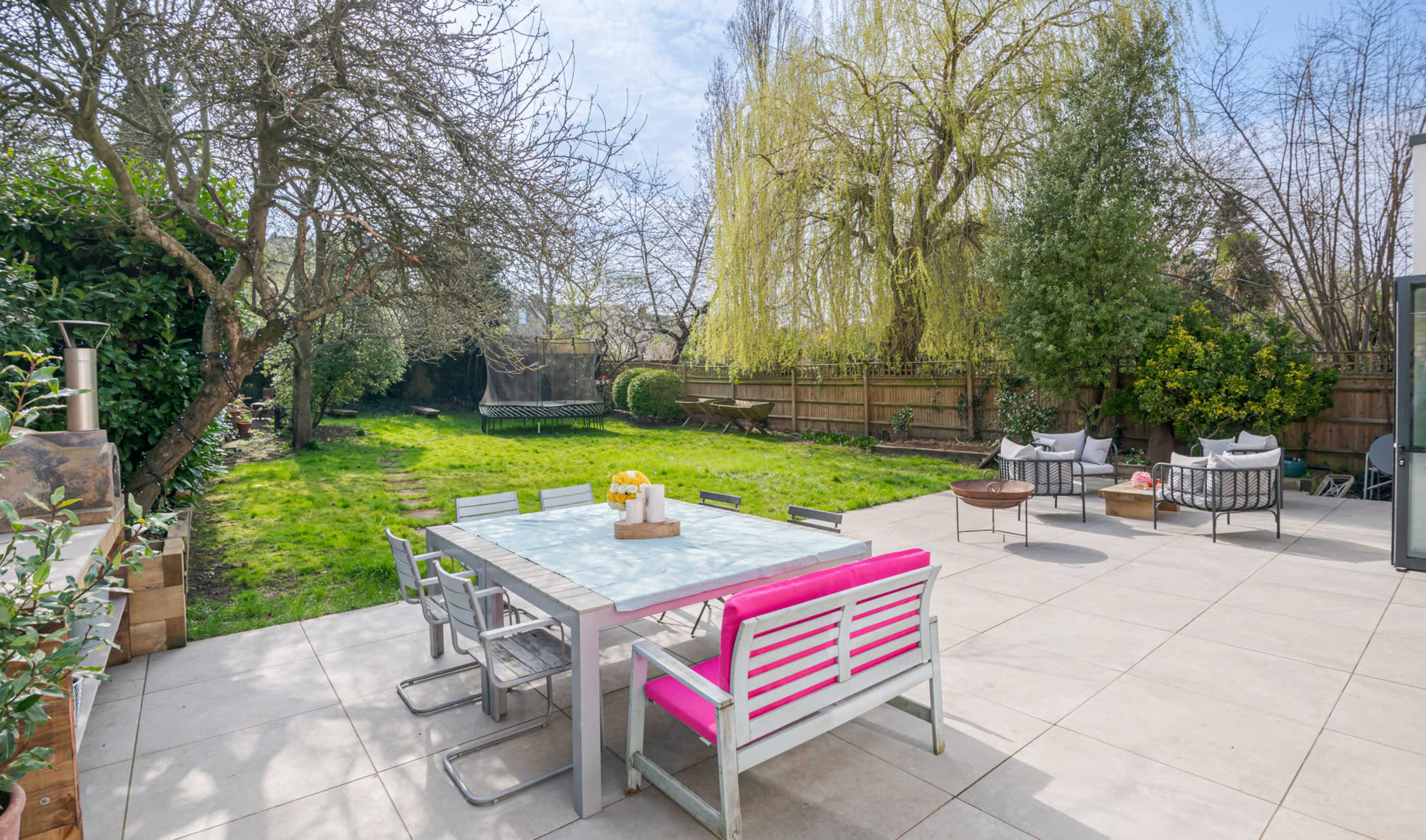 A patio with a pink-accented dining table and chairs overlooks a lawn bordered by trees and shrubs.