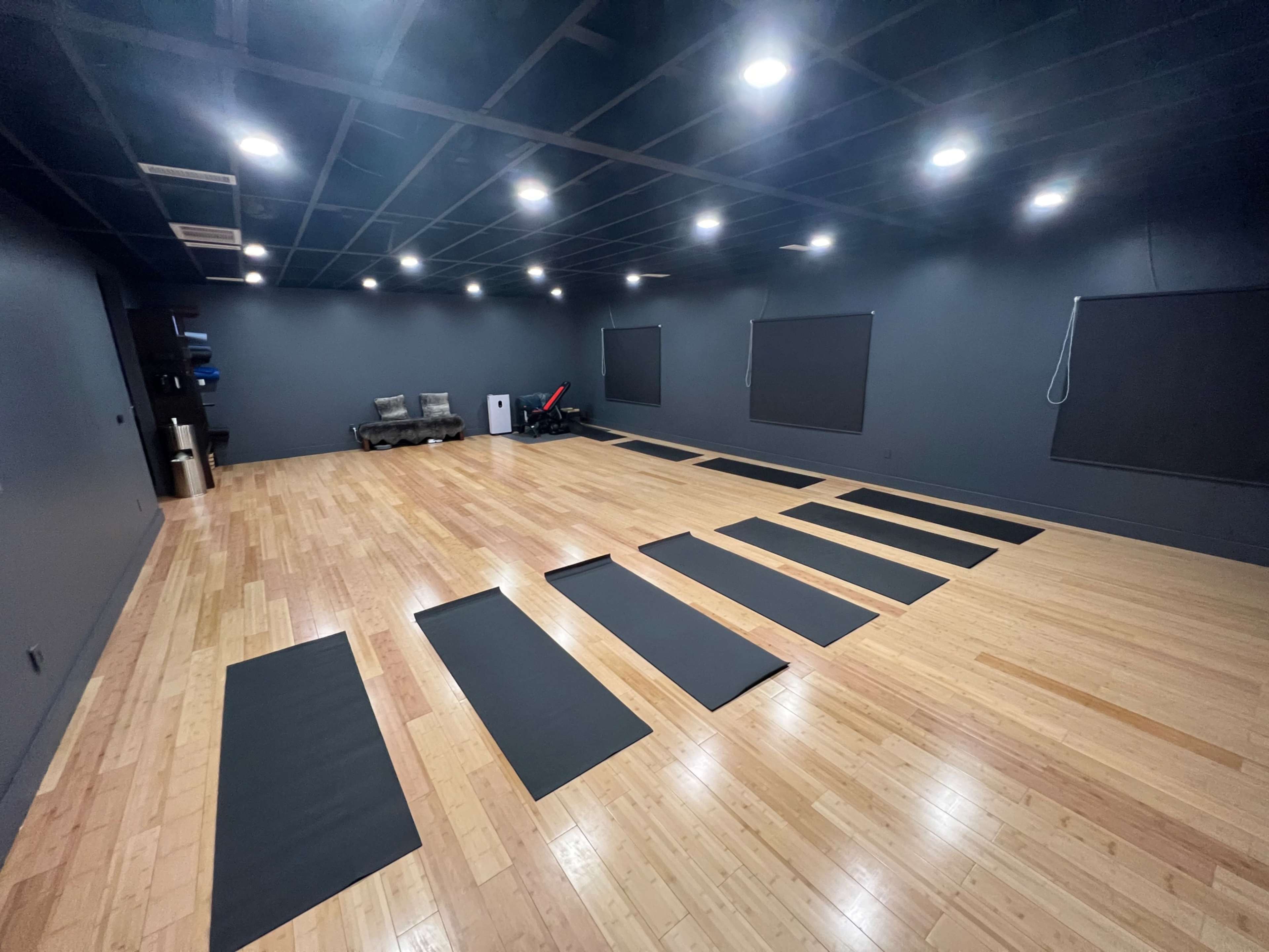 The image shows a spacious yoga or fitness studio with wooden flooring and several dark yoga mats arranged in rows against a backdrop of black walls and dim lighting.