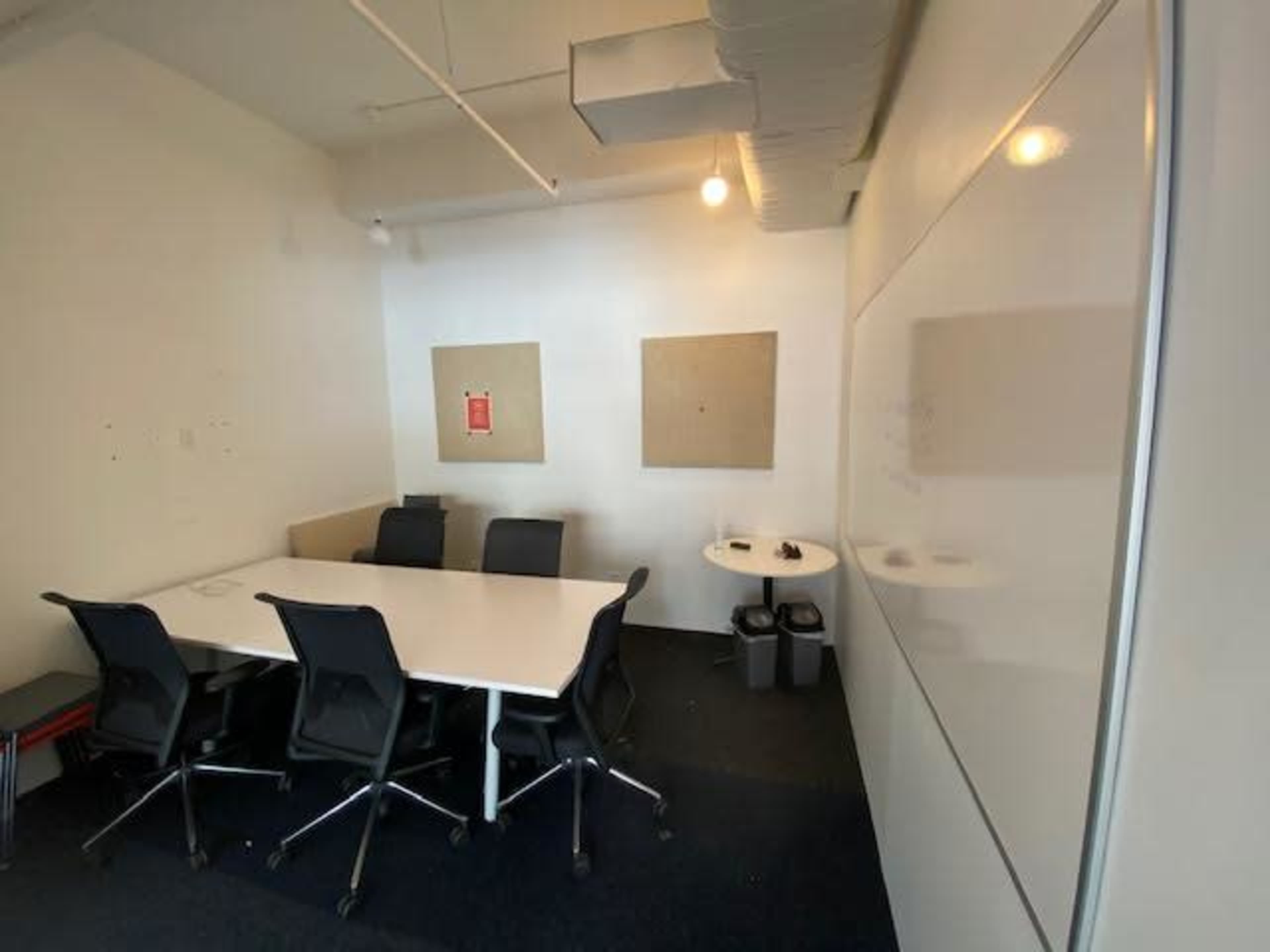 A conference room with a large white table surrounded by black chairs, two blank bulletin boards on the walls, and a small trash bin in the corner.