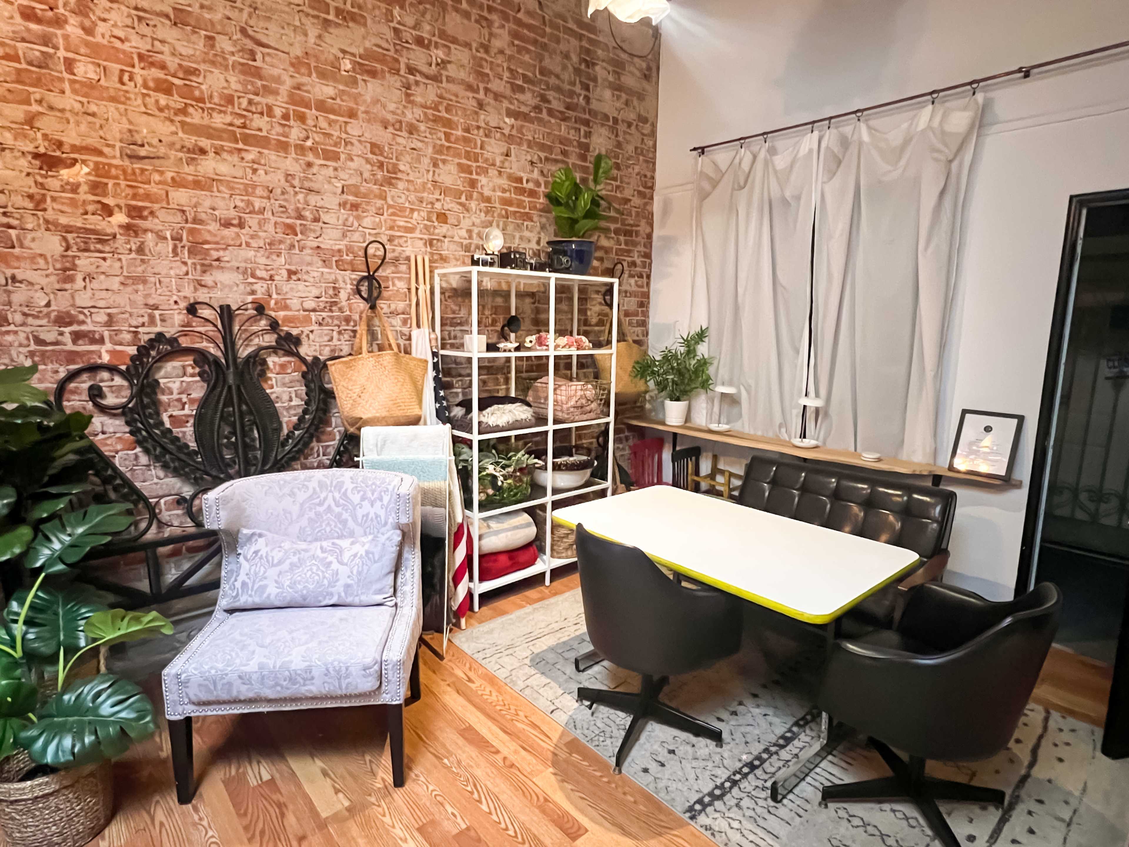 A cozy interior space featuring a brick wall, a white shelving unit with various items, two distinct chairs, a table with a yellow edge, and decorative plants.