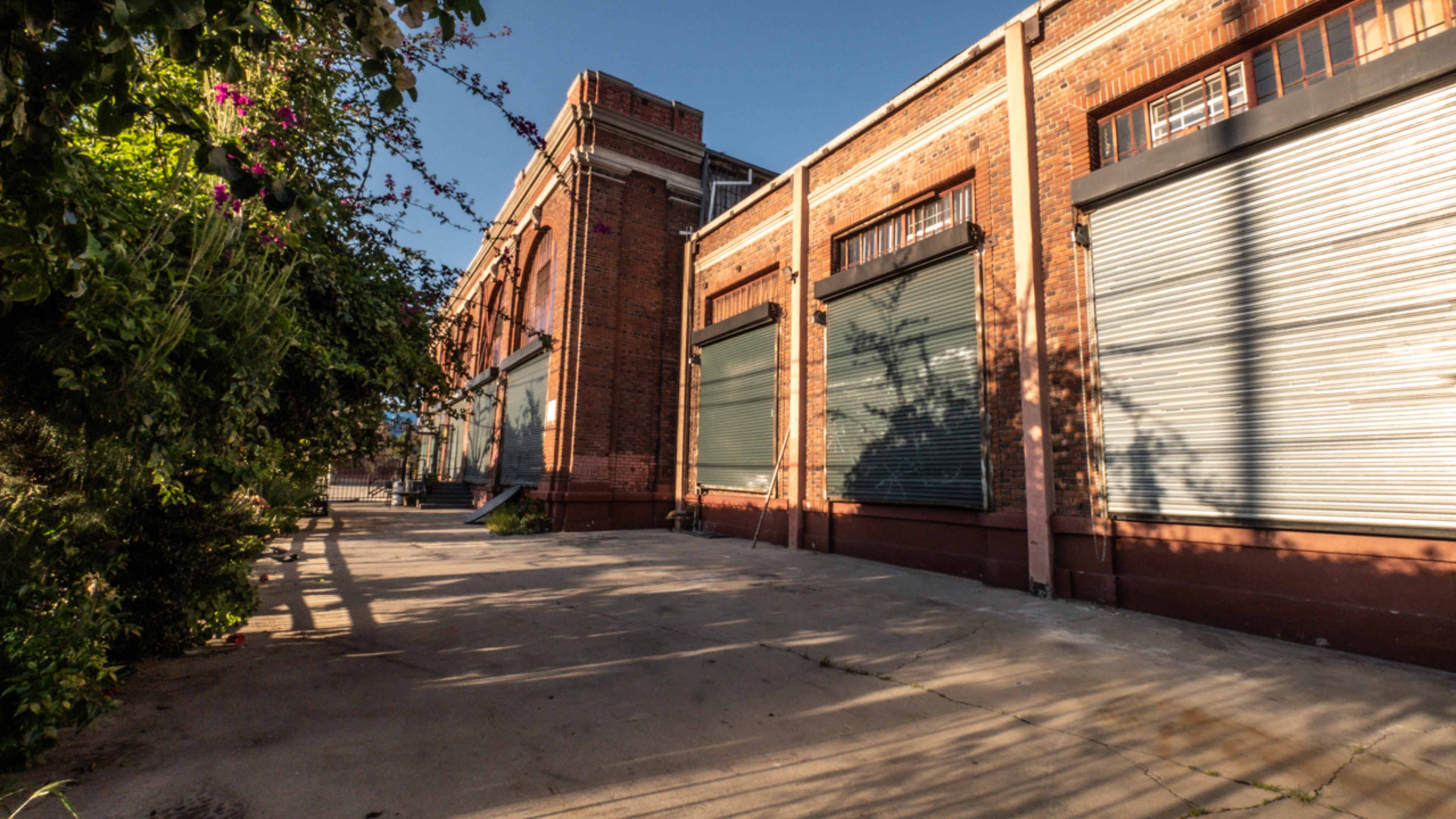 The image shows a brick building with closed metal shutters beside a paved walkway lined with greenery.