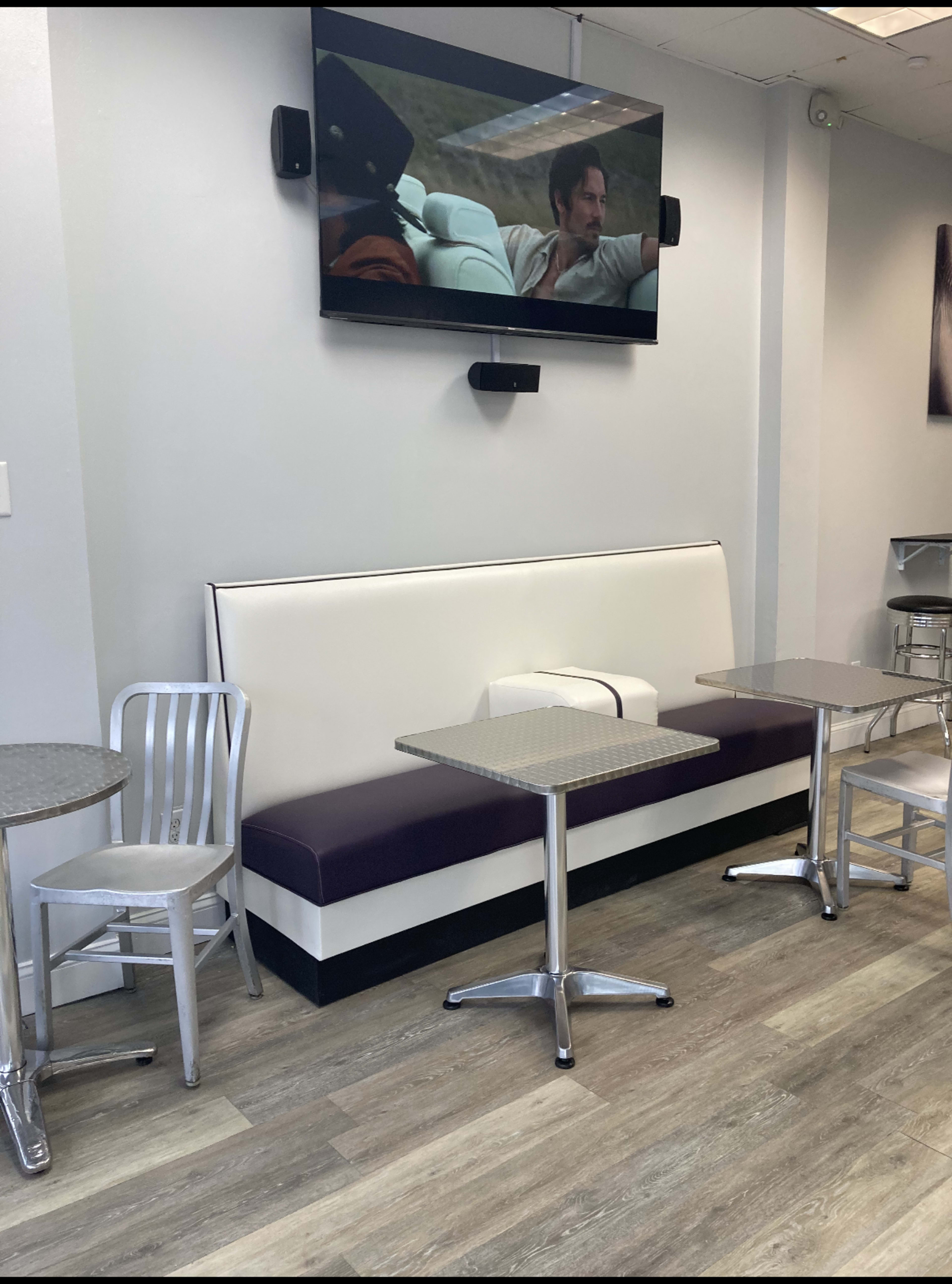 The image shows a minimalist cafe interior with a white and purple bench, two silver tables, and chairs arranged around them, along with a television mounted on the wall displaying a scene from a movie.