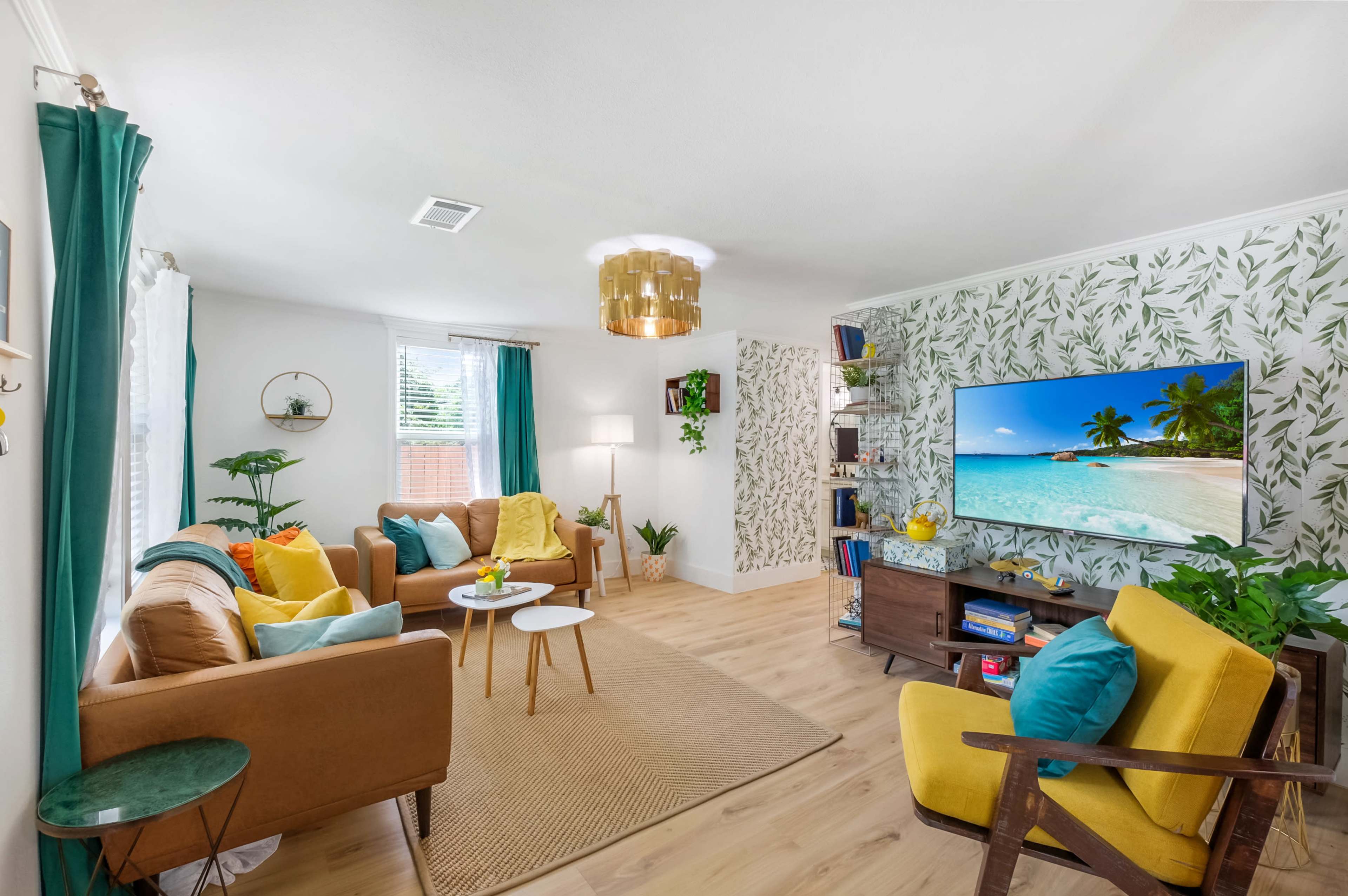 The living room features a combination of brown and yellow furniture, a large television mounted on a tropical-themed wall, and various plants decoratively arranged around the space.