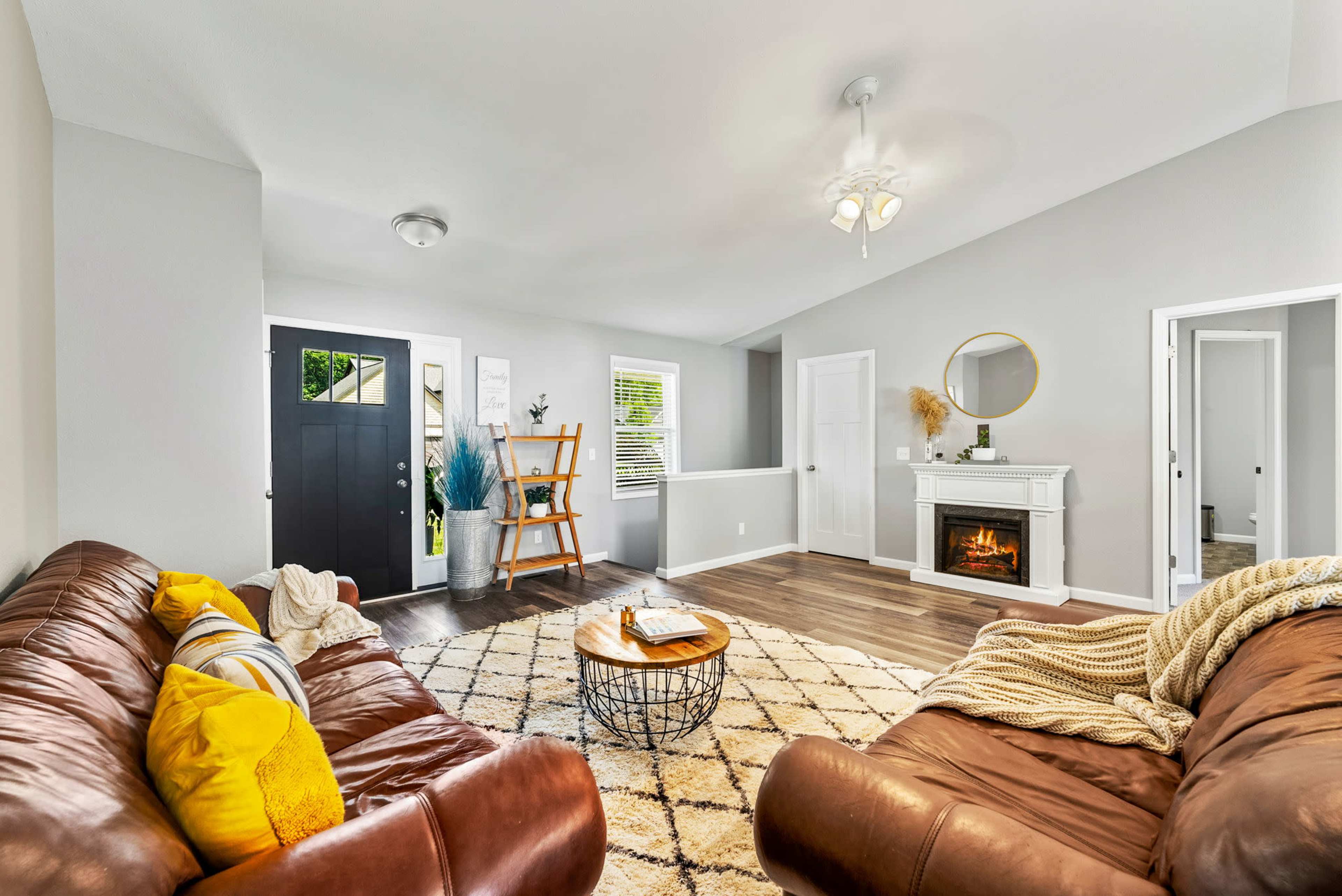 A modern living room features two brown leather sofas, a round wooden coffee table, and a faux fireplace, with a black front door and large mirror visible in the background.