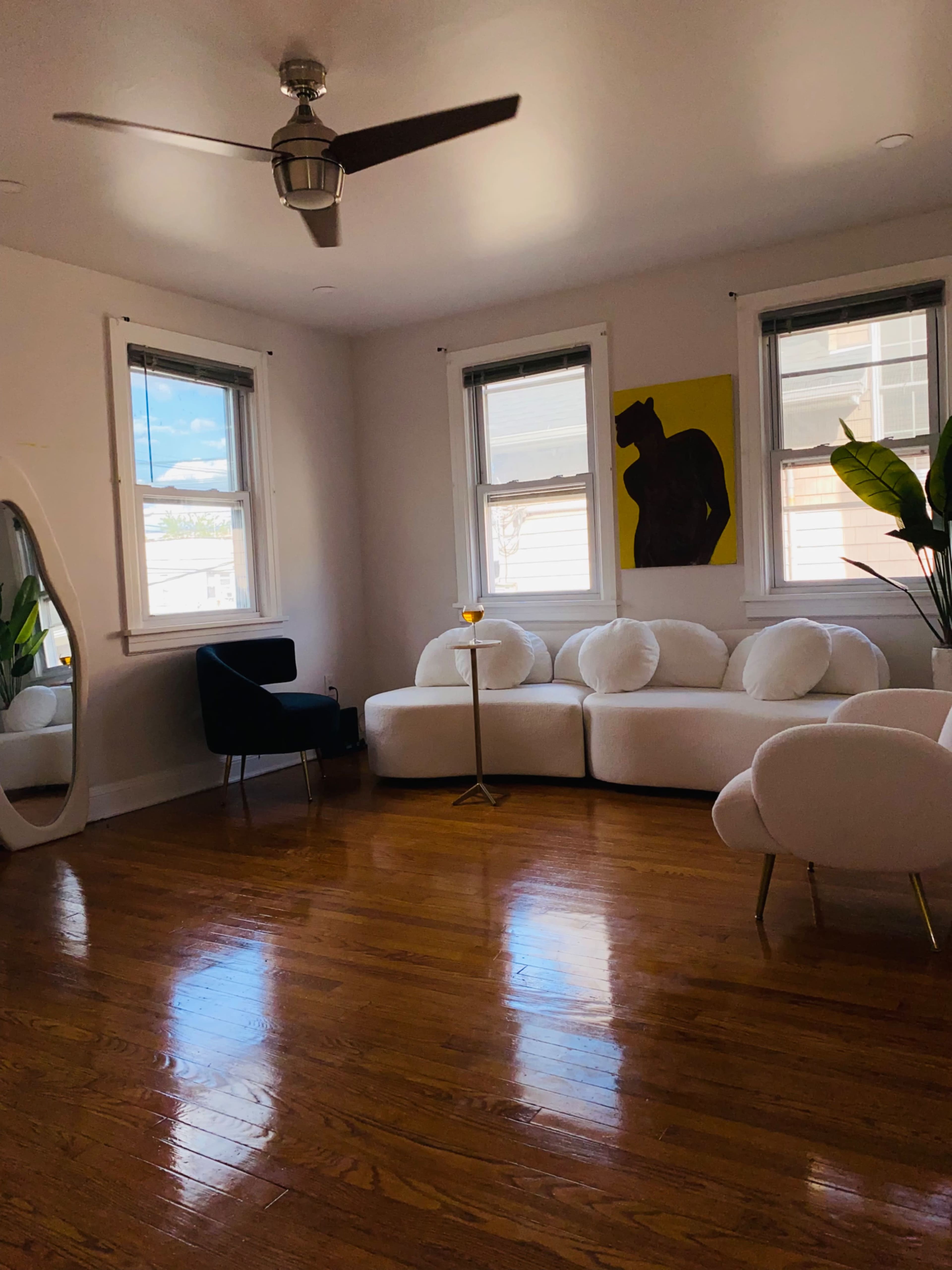 A spacious living room features a large white sofa, a black chair, a tall mirror, and a plant, with natural light coming through two windows.