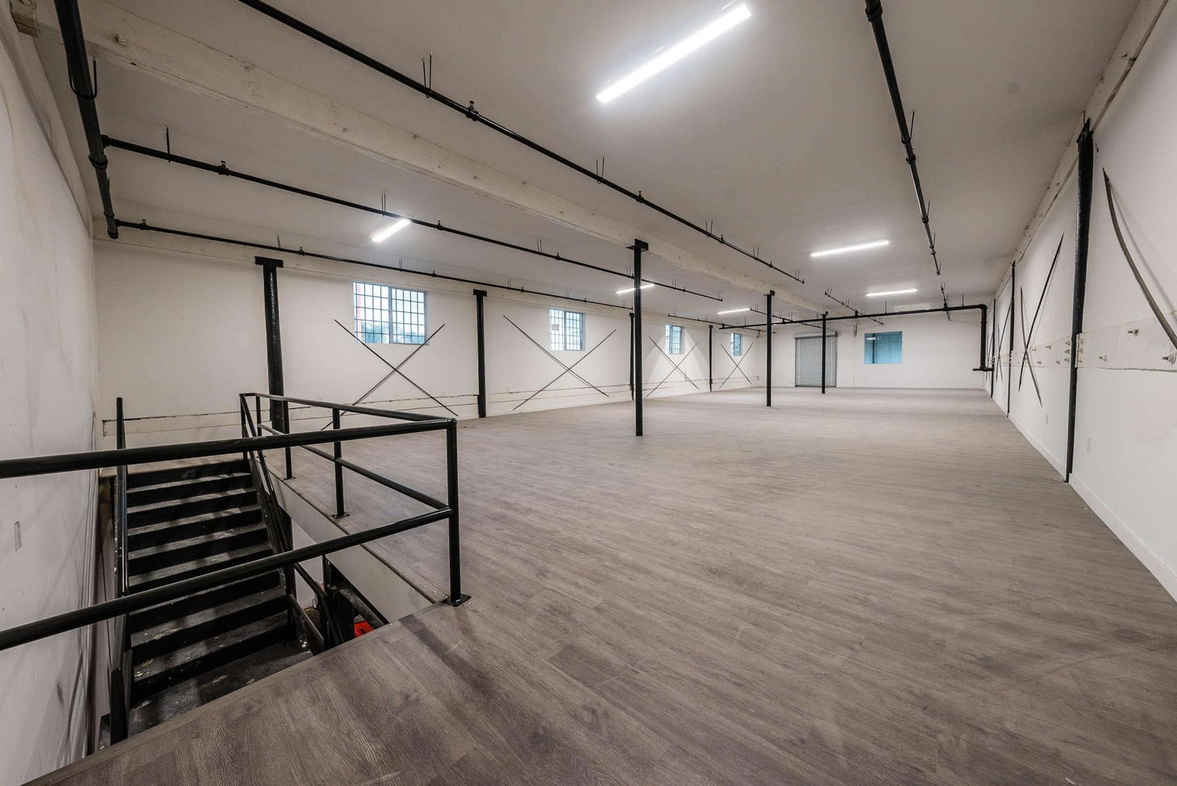 The image shows a spacious, empty room with light-colored walls, a wooden floor, and large windows.