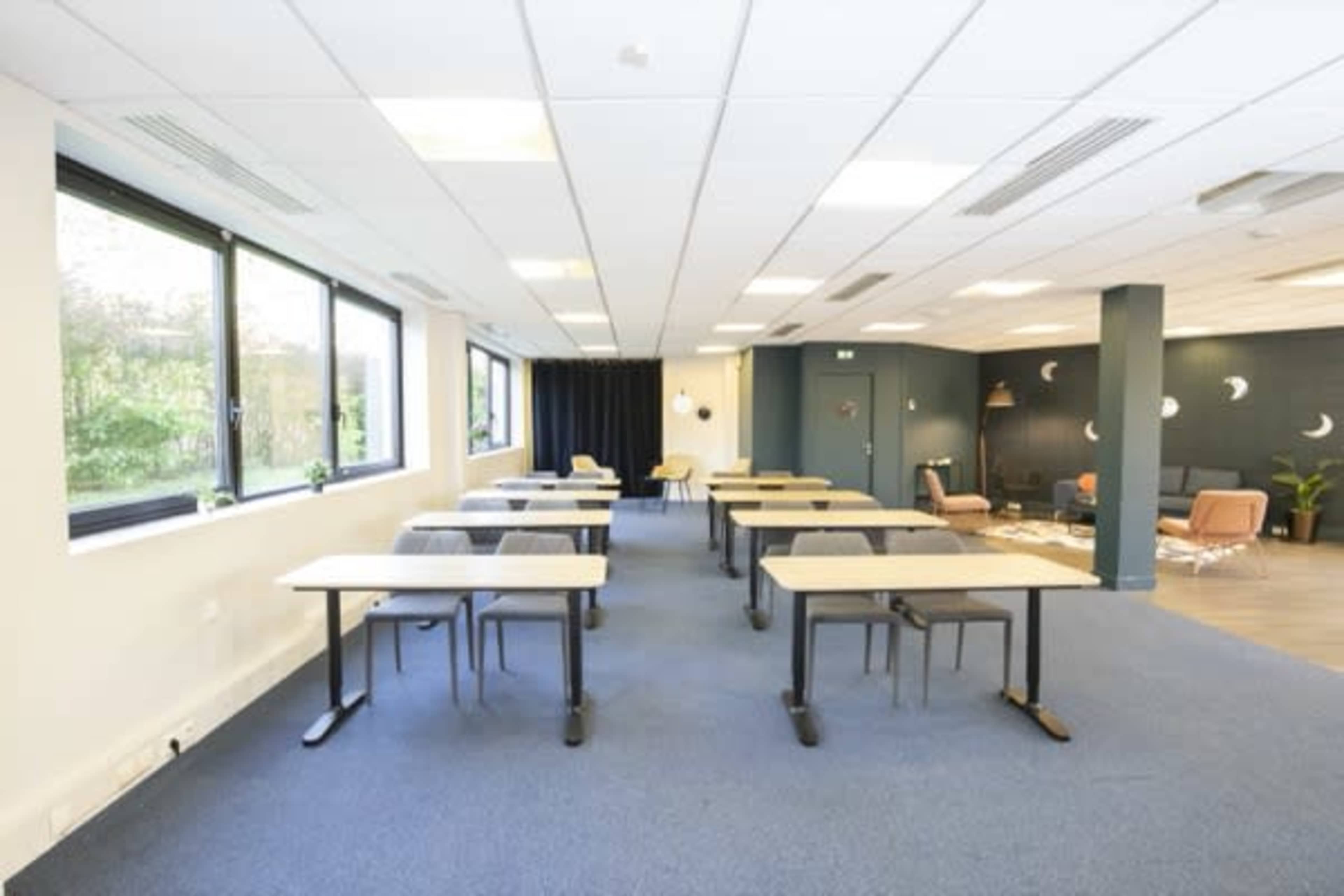 A spacious classroom or meeting room with several rows of tables and chairs arranged neatly, featuring large windows that let in natural light.