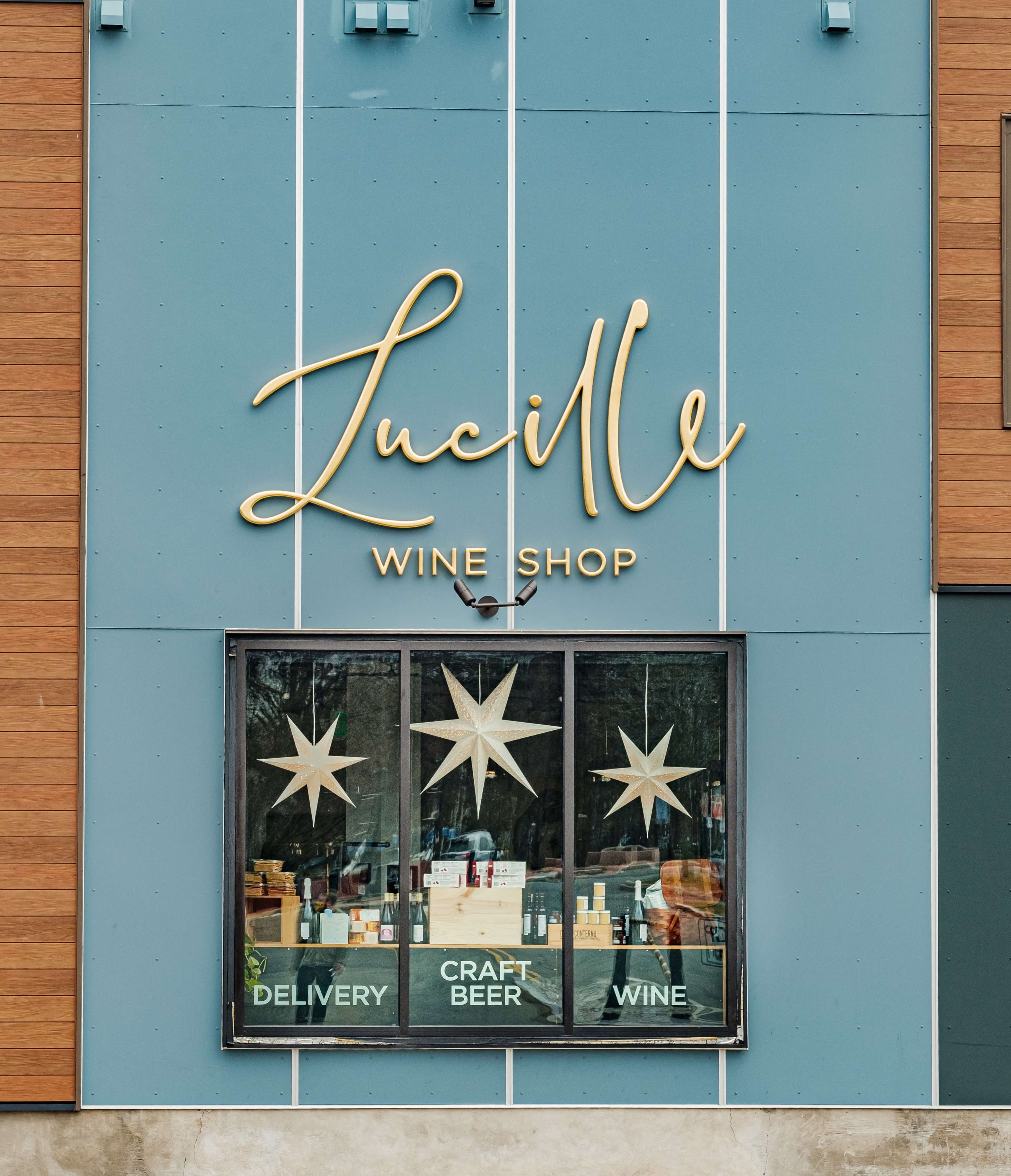 A storefront of "Lucille Wine Shop," featuring a large sign and a window displaying promotions for delivery, craft beer, and wine.